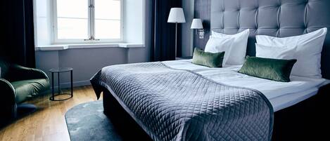 A hotel room with a large bed, a bedside table, a lamp, and a view of the outdoors through a window.