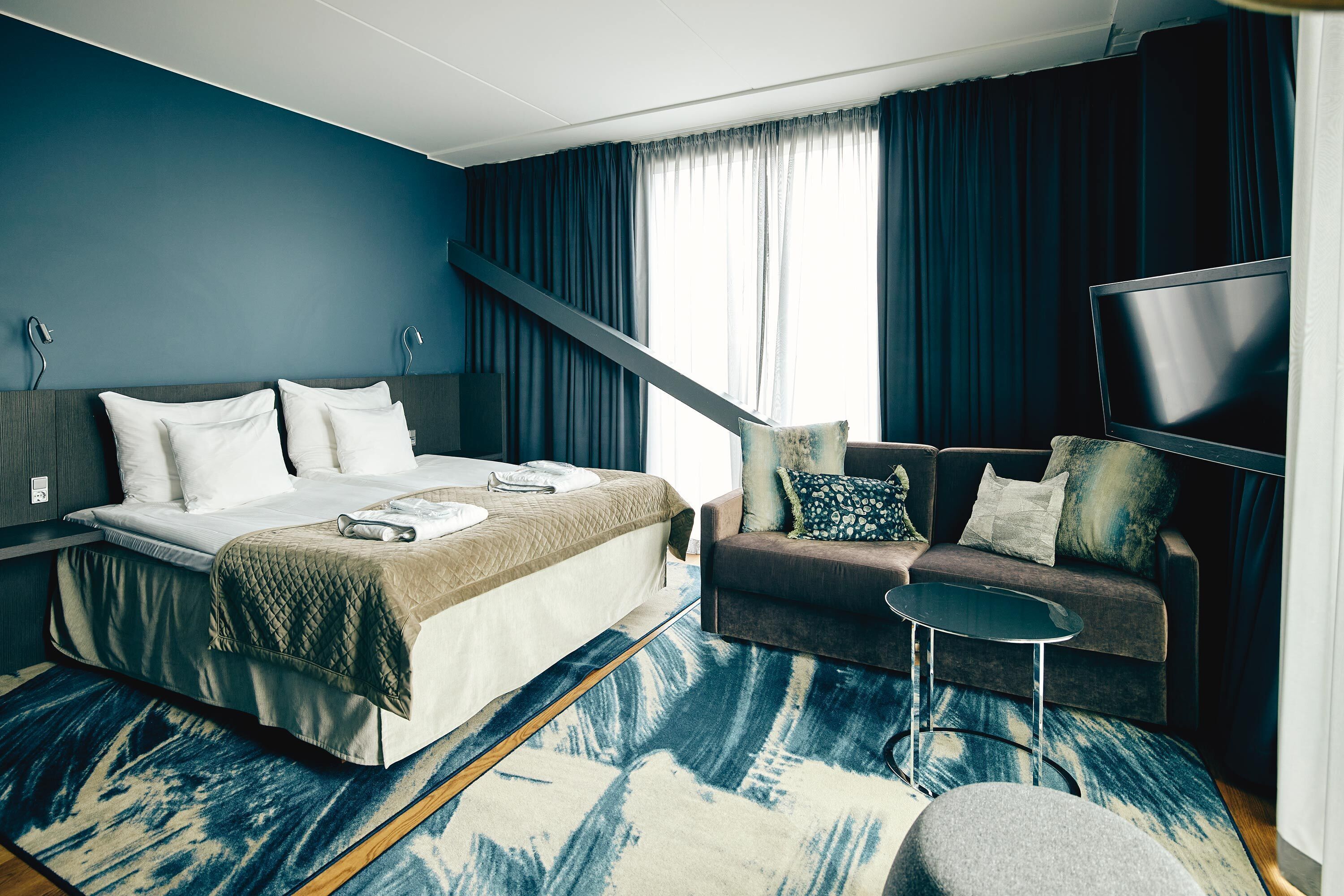 Et moderne hotelværelse med seng, sofa, fjernsyn og et stort vindue med gardiner.