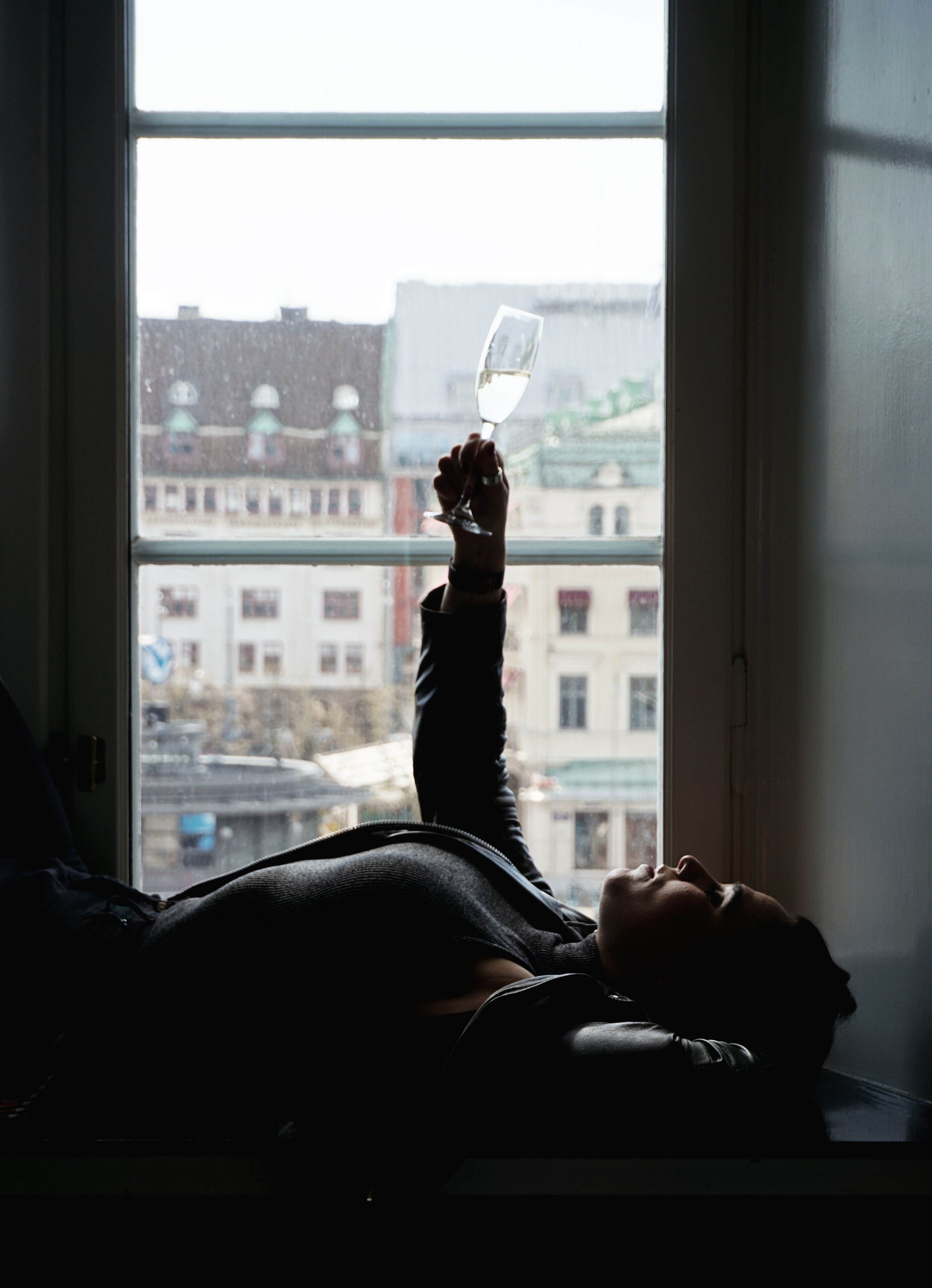 A person lying on a bed by a window with a view of buildings outside.