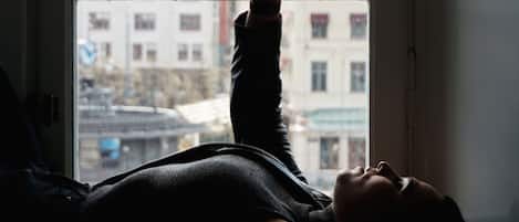 A person lying on a bed by a window with a view of buildings outside.