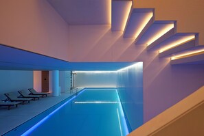A modern indoor swimming pool with a minimalist design, featuring a long, narrow pool and a series of steps leading down to the water.