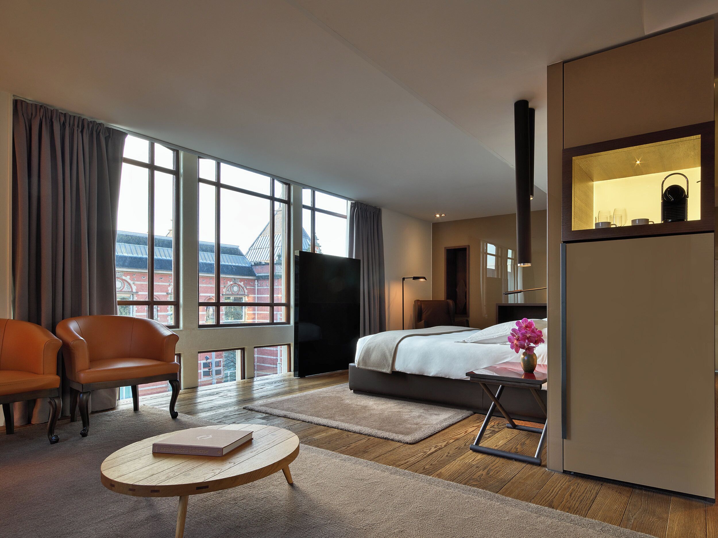 Ett hotellrum med en stor säng, två stolar, ett litet bord och utsikt över byggnader genom stora fönster.