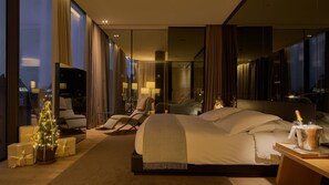 Un dormitorio de hotel moderno con una cama grande, un área de descanso con sillas, un pequeño árbol de Navidad y una mesita de noche con una botella de champán.