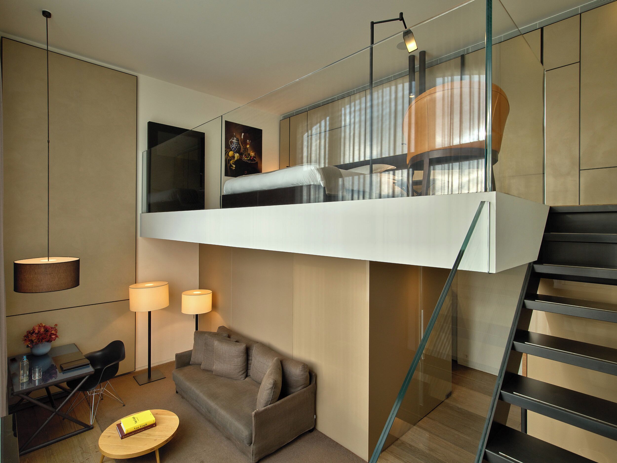 Une chambre moderne de style loft, dotée d’un escalier en verre, d’un canapé, d’une petite table et d’un bureau avec une lampe.