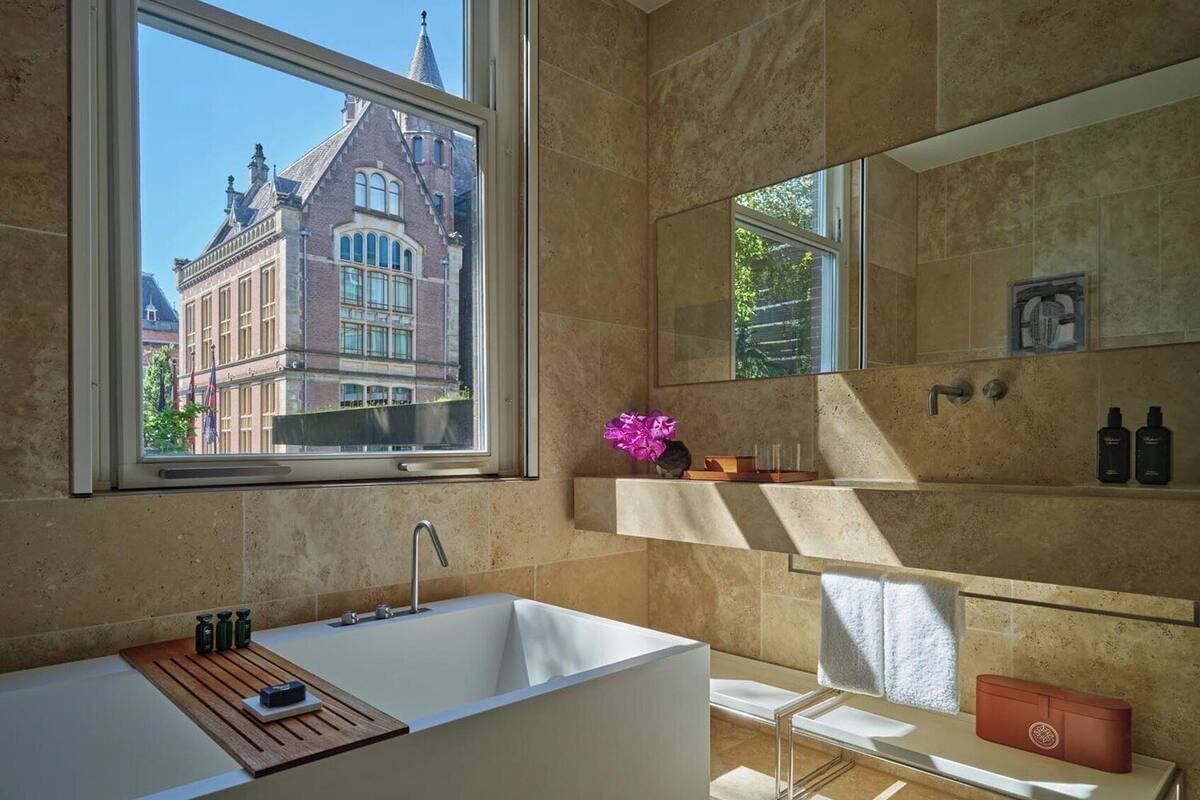 Ein modernes Badezimmer mit einer großen Badewanne, einem Fenster mit Blick auf ein historisches Gebäude, einem Spiegel und einem Ablagefach mit Pflegeprodukten.