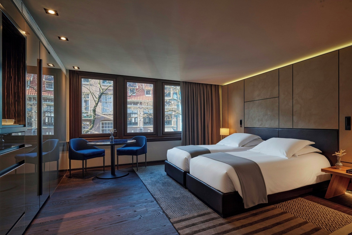 Une chambre d’hôtel avec un lit, deux chaises, un bureau, une lampe et de grandes fenêtres.