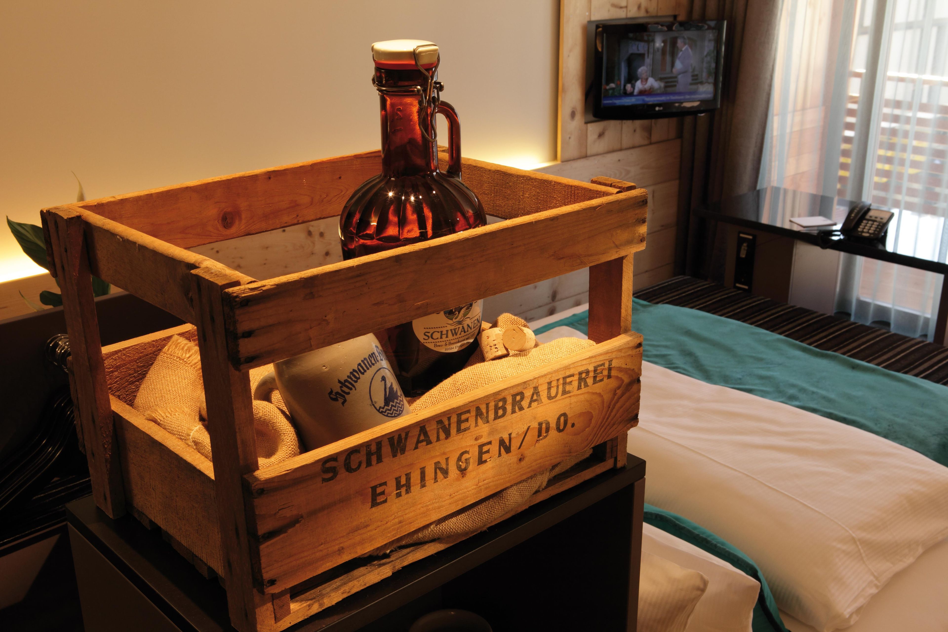 A wooden crate with a bottle and a bottle of Schweppes, placed on a bed with a view of a TV in the background.
