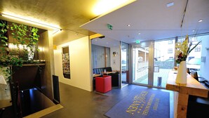 Een moderne hotellobby met een receptiebalie, een rode zithoek en een groot raam met uitzicht op de straat.