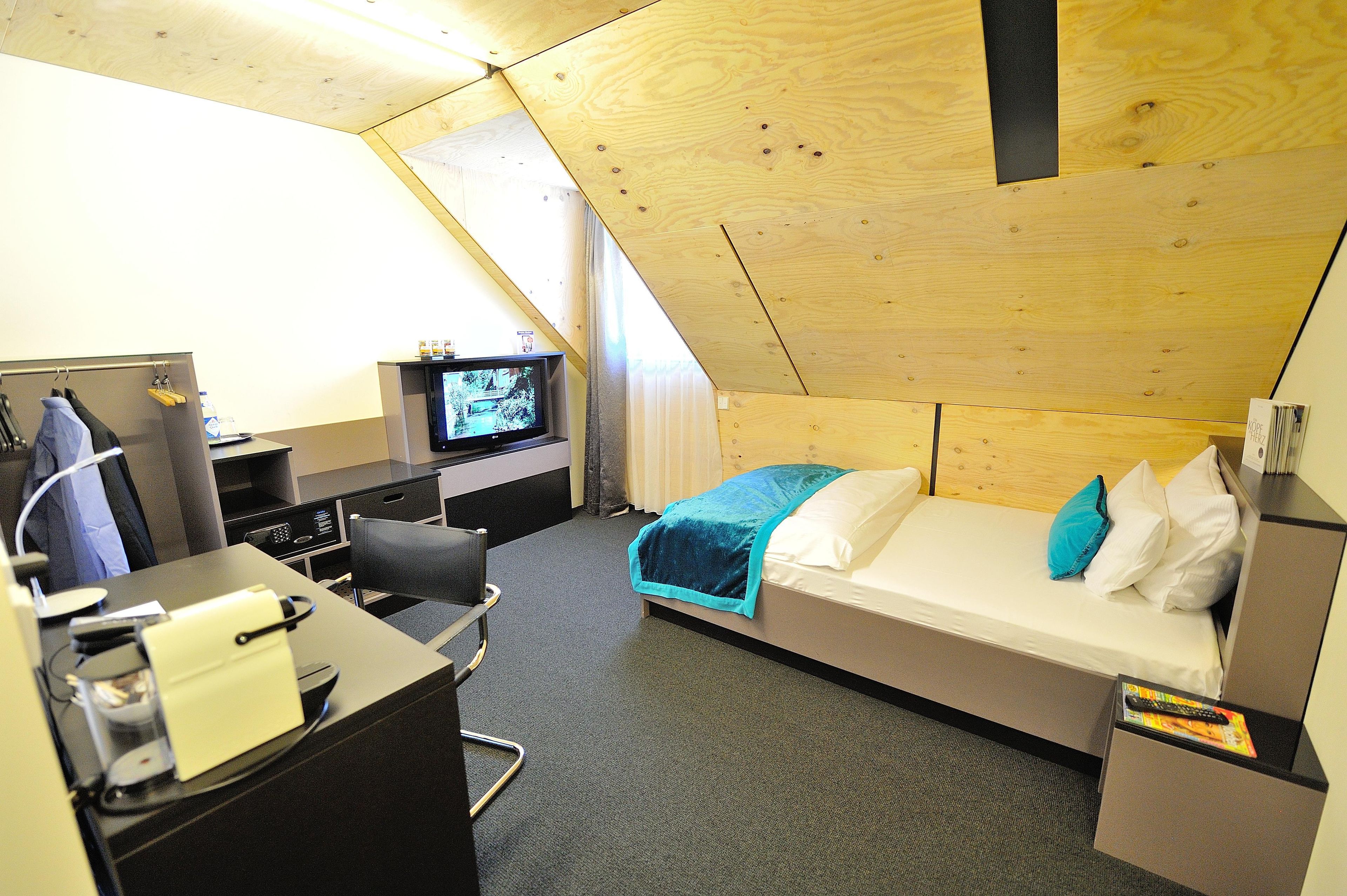 A small, triangular room with a bed, a desk with a computer, and a television.