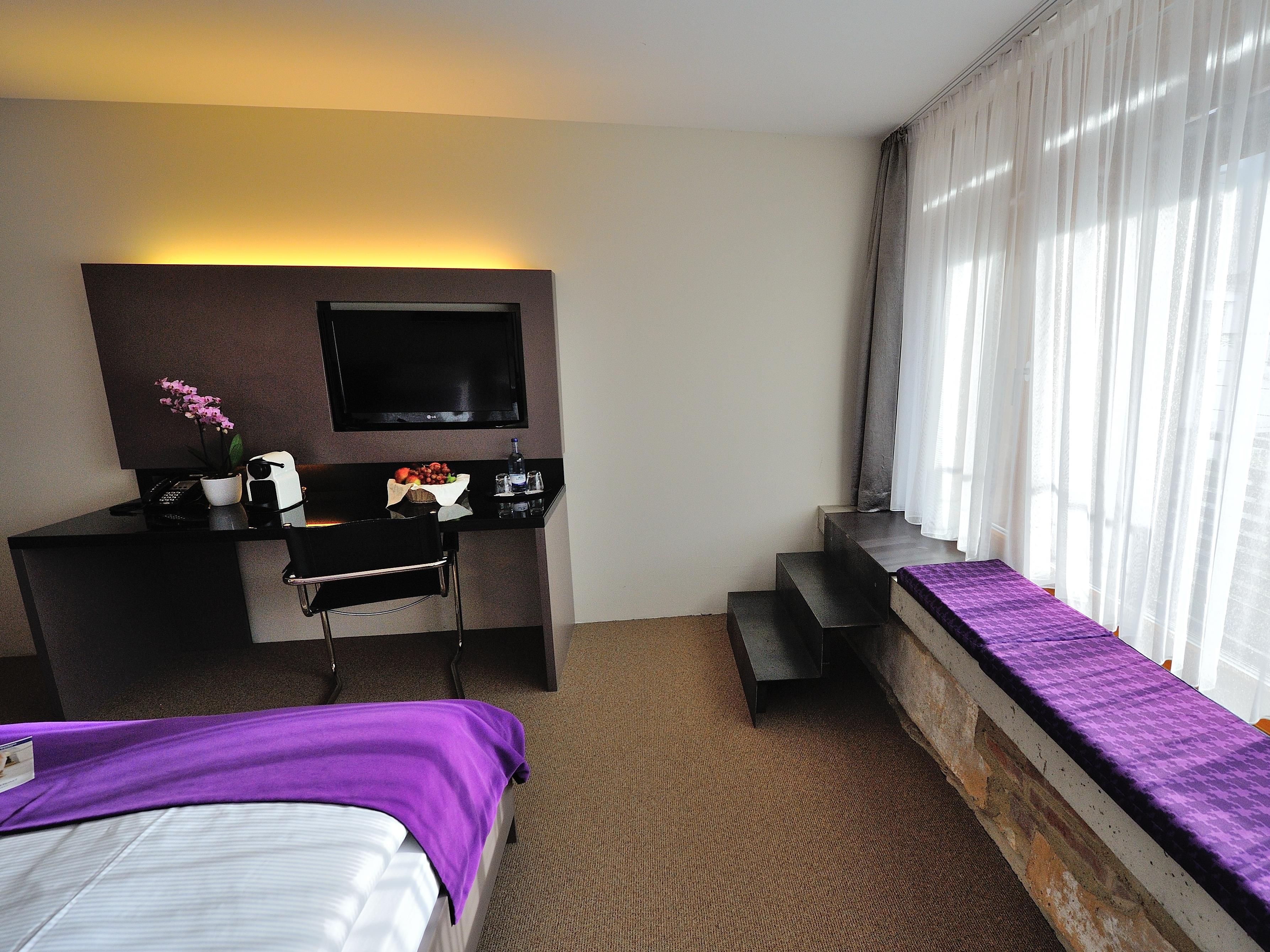 A hotel room with a bed, a desk with a computer and a flat-screen TV, a small table with fruits and a bottle, and a window with curtains.