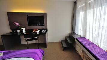 Quarto de hotel com cama, escrivaninha com computador e TV de tela plana, mesinha com frutas e uma garrafa, e janela com cortinas.