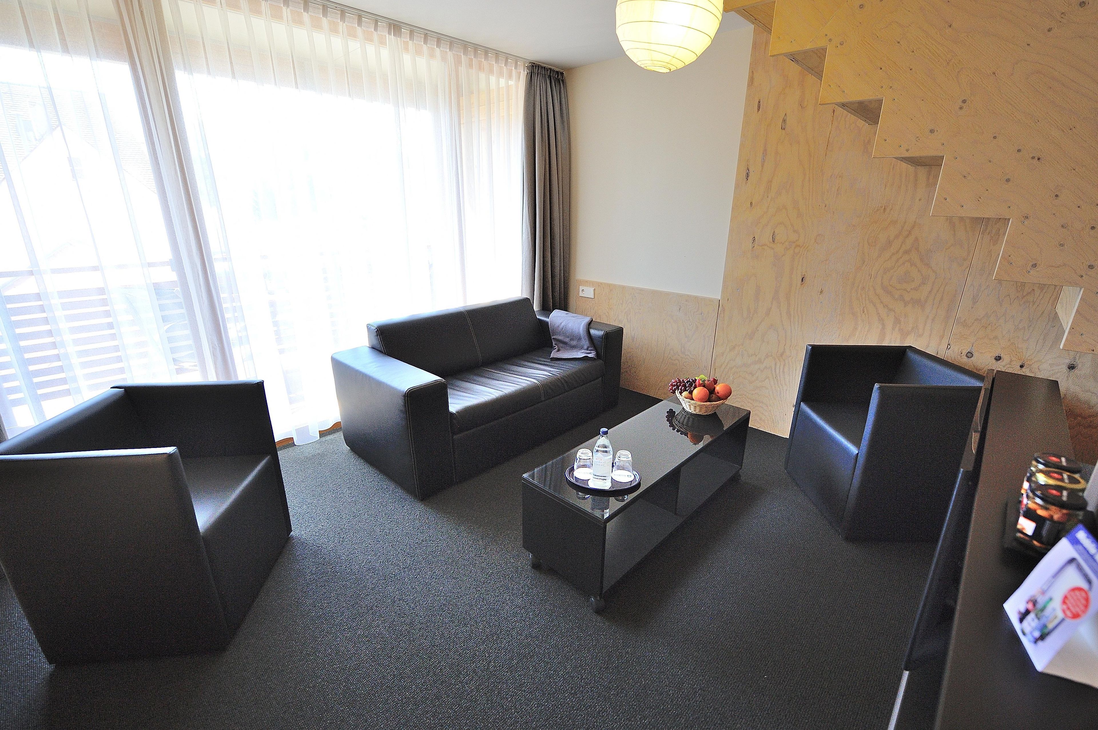 A modern hotel room with a sofa, two armchairs, a coffee table with a fruit bowl, and a wooden accent wall.