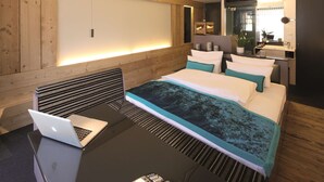 Moderni hotellihuone, jossa on puukoristeinen seinä, turkoosinsävyisillä tekstiileillä varustettu sänky, työpöytä kannettavalla tietokoneella ja yöpöytä, jossa on kaukosäädin.