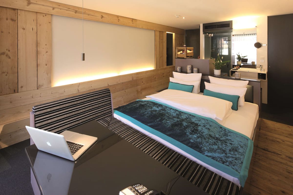 Une chambre d’hôtel moderne avec un mur d’accent en bois, un lit avec une literie de couleur turquoise, un bureau avec un ordinateur portable et une table de chevet avec une télécommande.
