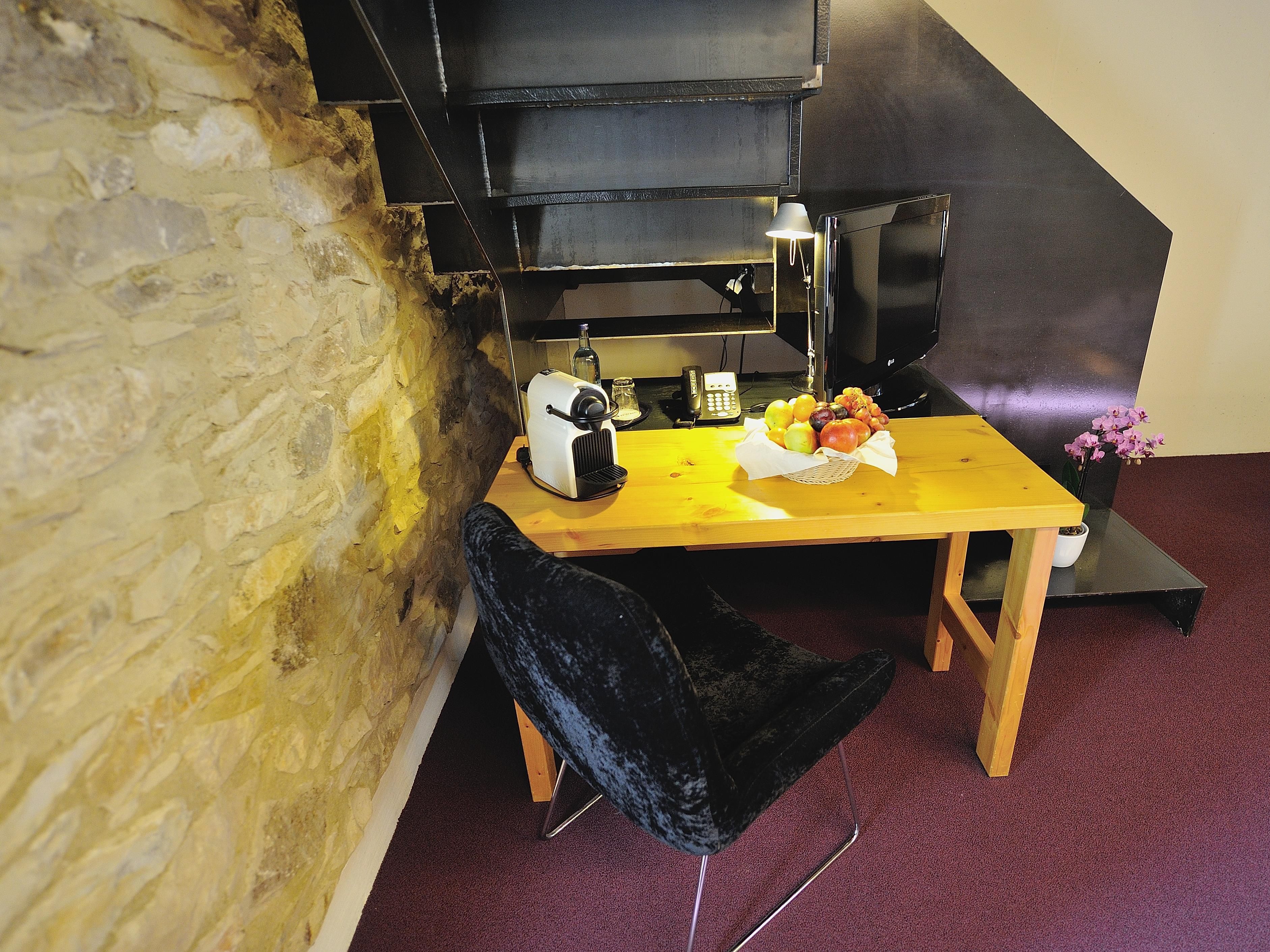 A room with a stone wall, a wooden table with a basket of fruit, a black chair, and a black grill in the background.