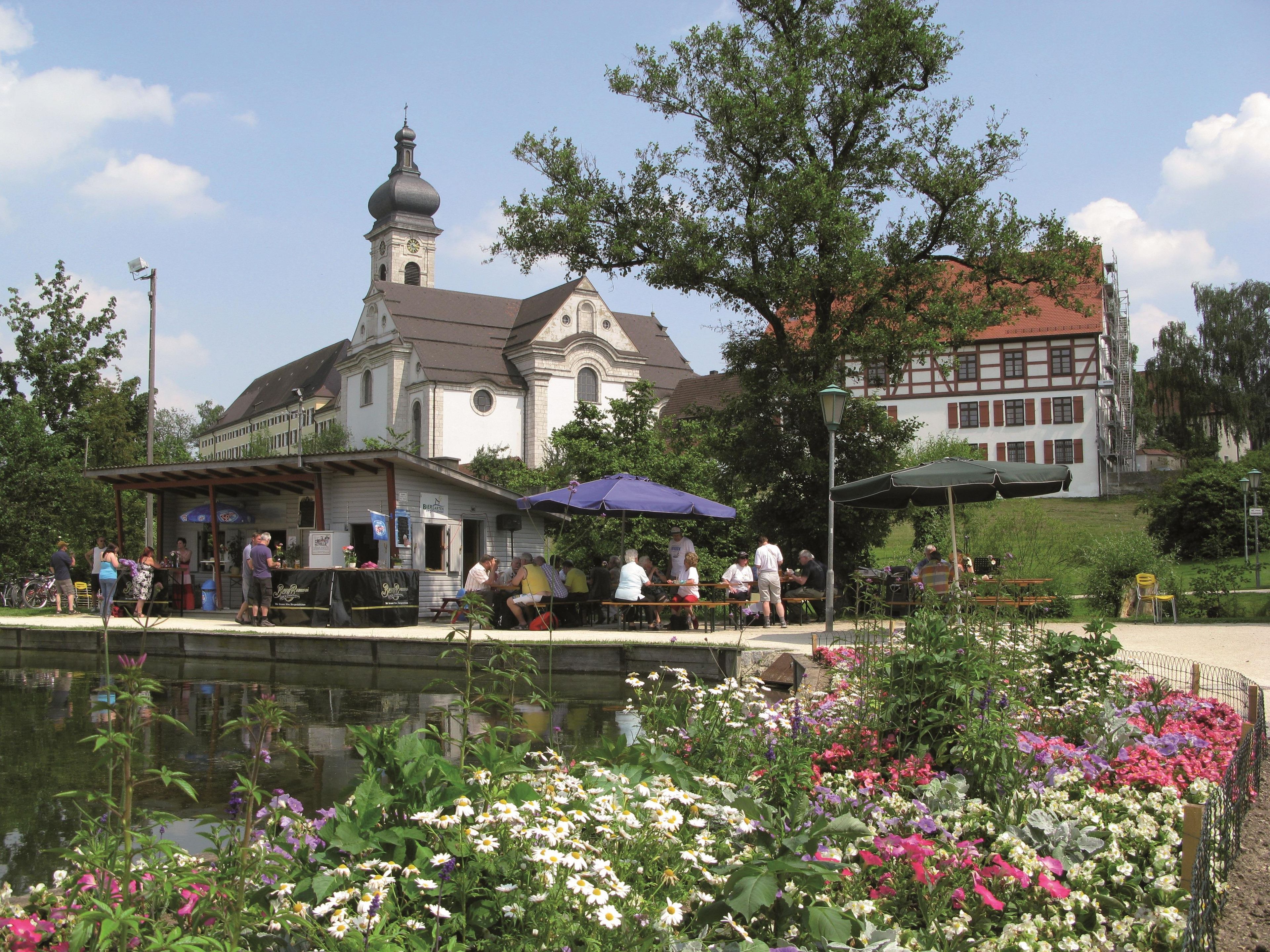 Eine Kirche mit einem Glockenturm, ein Teich mit Blumen und ein Gebäude mit rotem Dach.