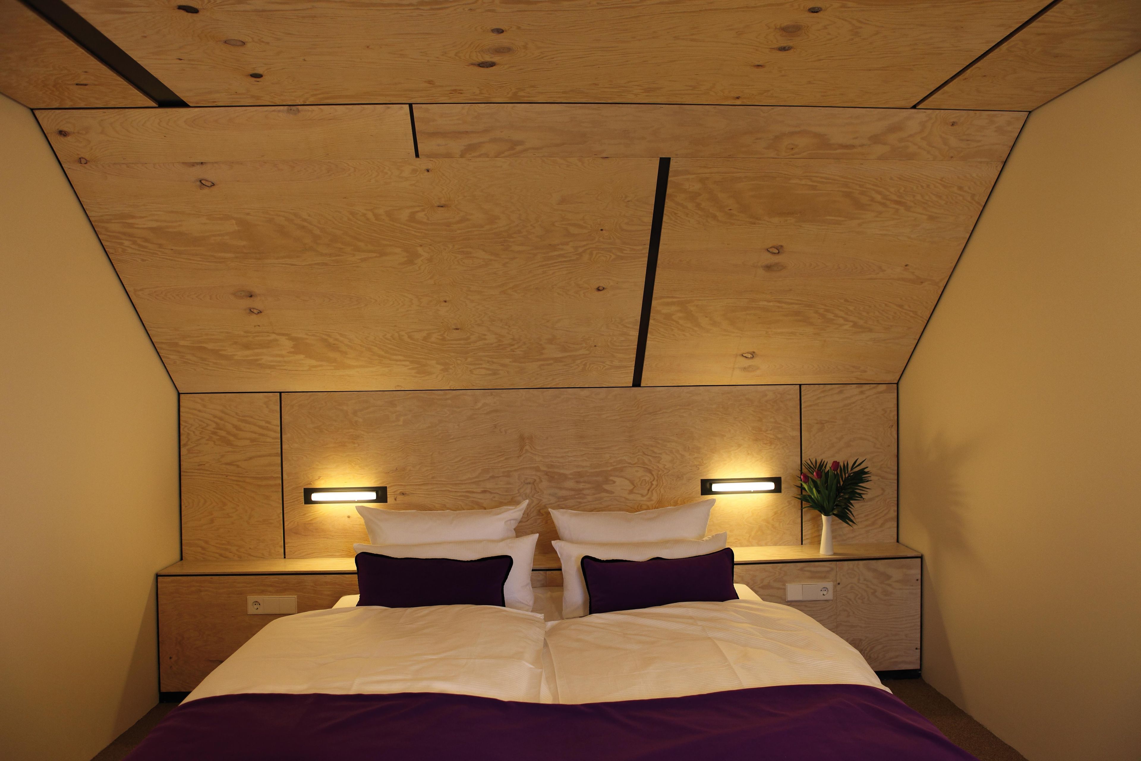 Une chambre d’hôtel avec un plafond en bois, deux lampes de chevet, un lit avec une literie blanche et violette, et une petite plante en pot posée sur la table de chevet.