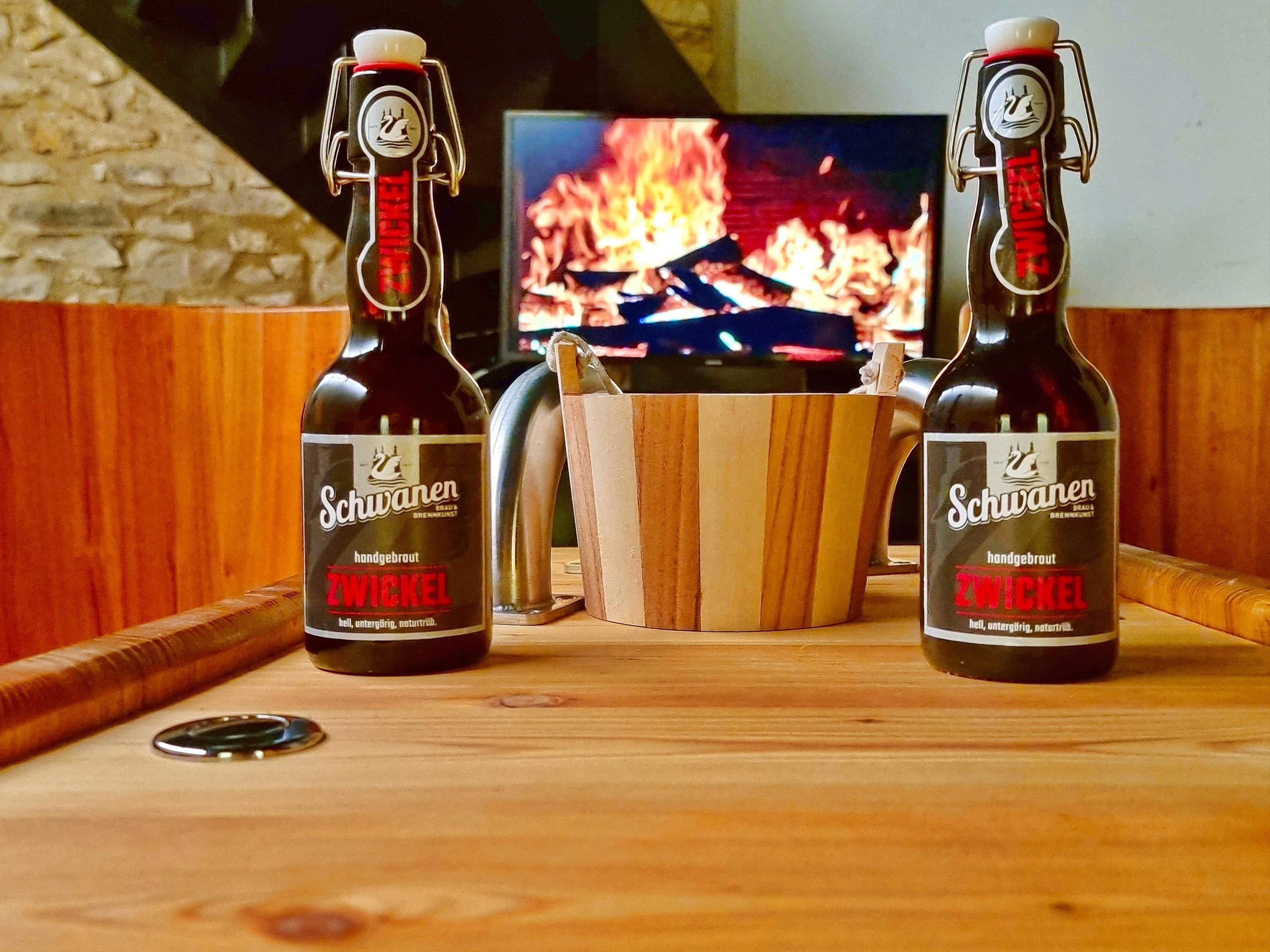 Zwei Flaschen Zwickelbier auf einer Holztischplatte, im Hintergrund ein Kaminfeuer.