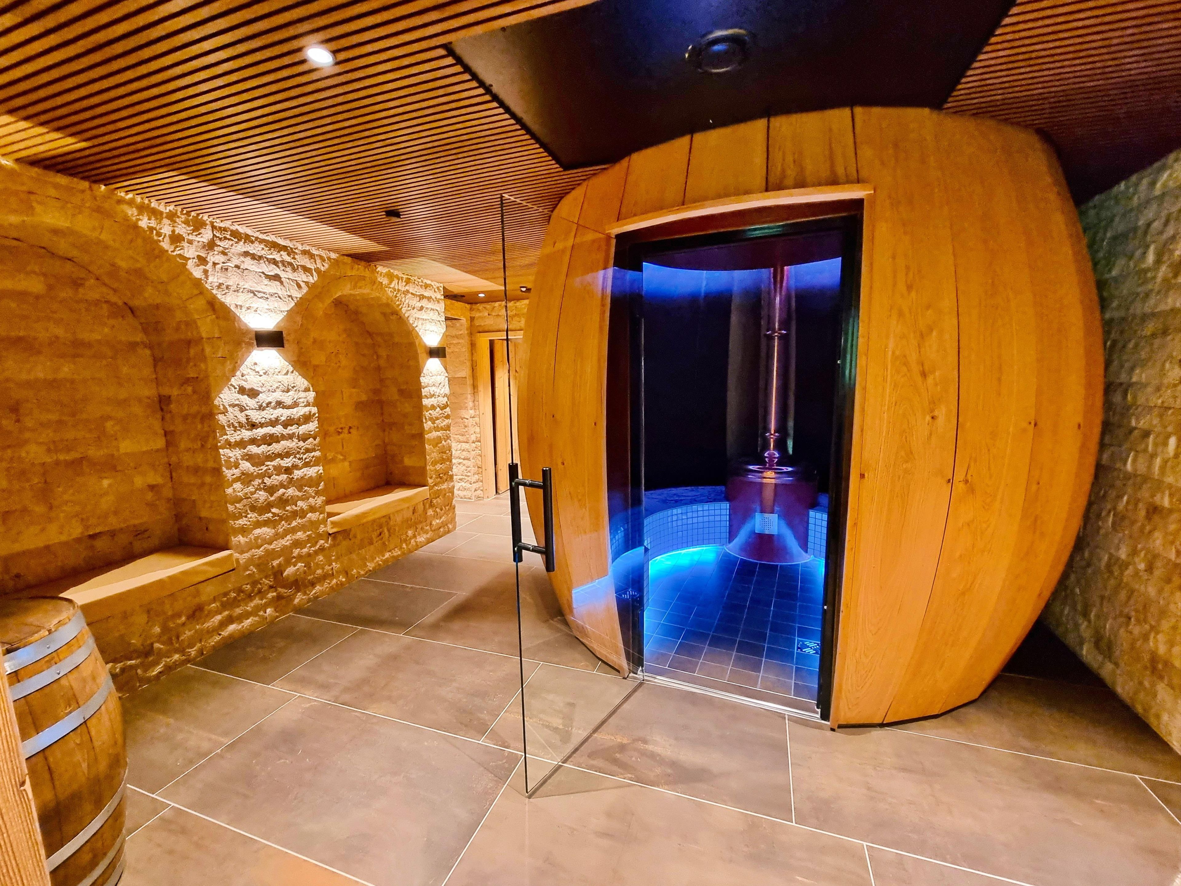 Ein Wellnessraum mit einem Holzfass, Steinwänden und einer blau beleuchteten Wasserspülung.