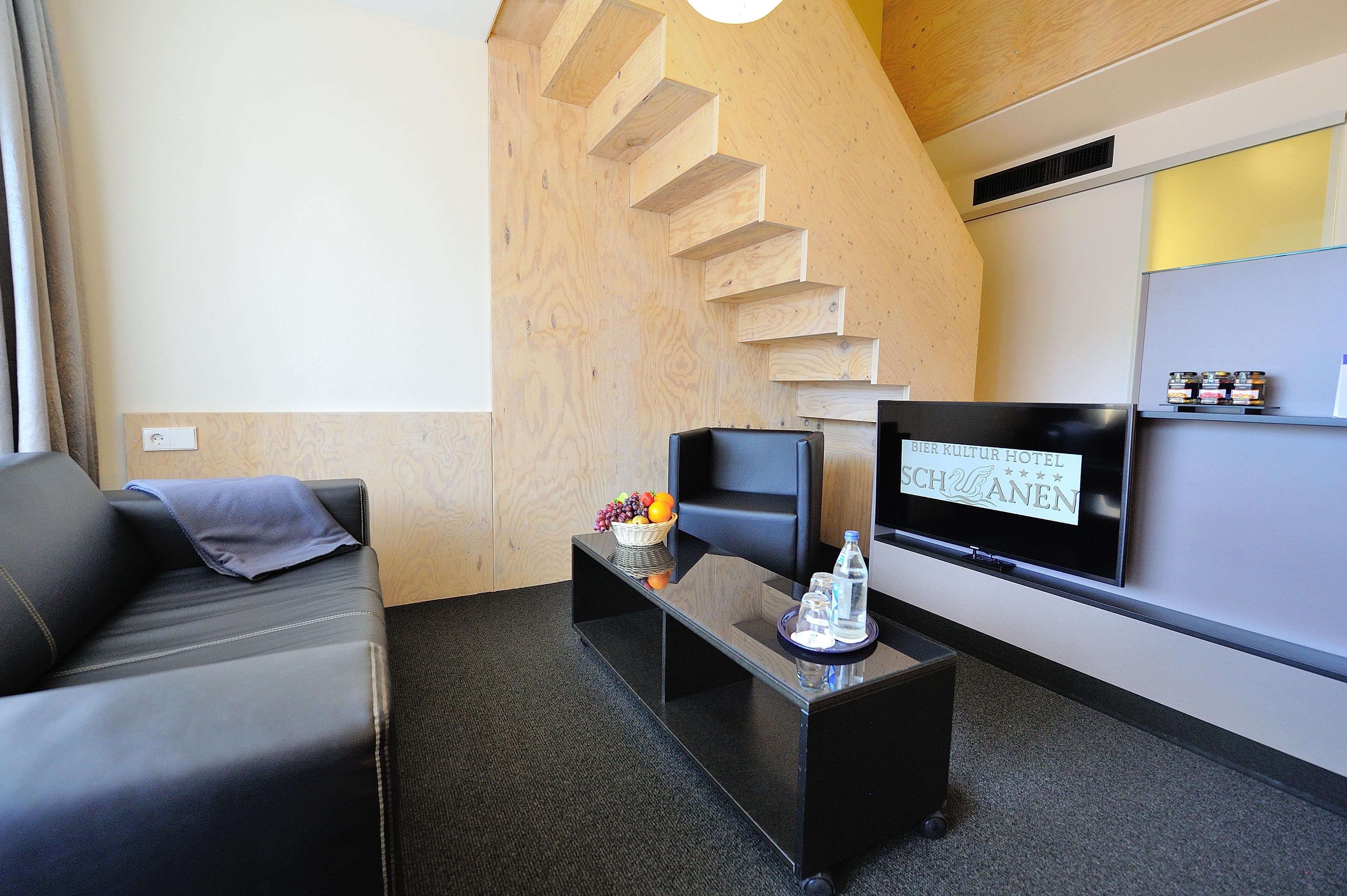 A hotel room with a modern design, featuring a sofa, a flat-screen TV, a small table with a fruit basket, and a wall-mounted shelf with items.