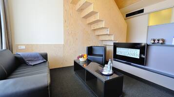 A hotel room with a modern design, featuring a sofa, a flat-screen TV, a small table with a fruit basket, and a wall-mounted shelf with items.
