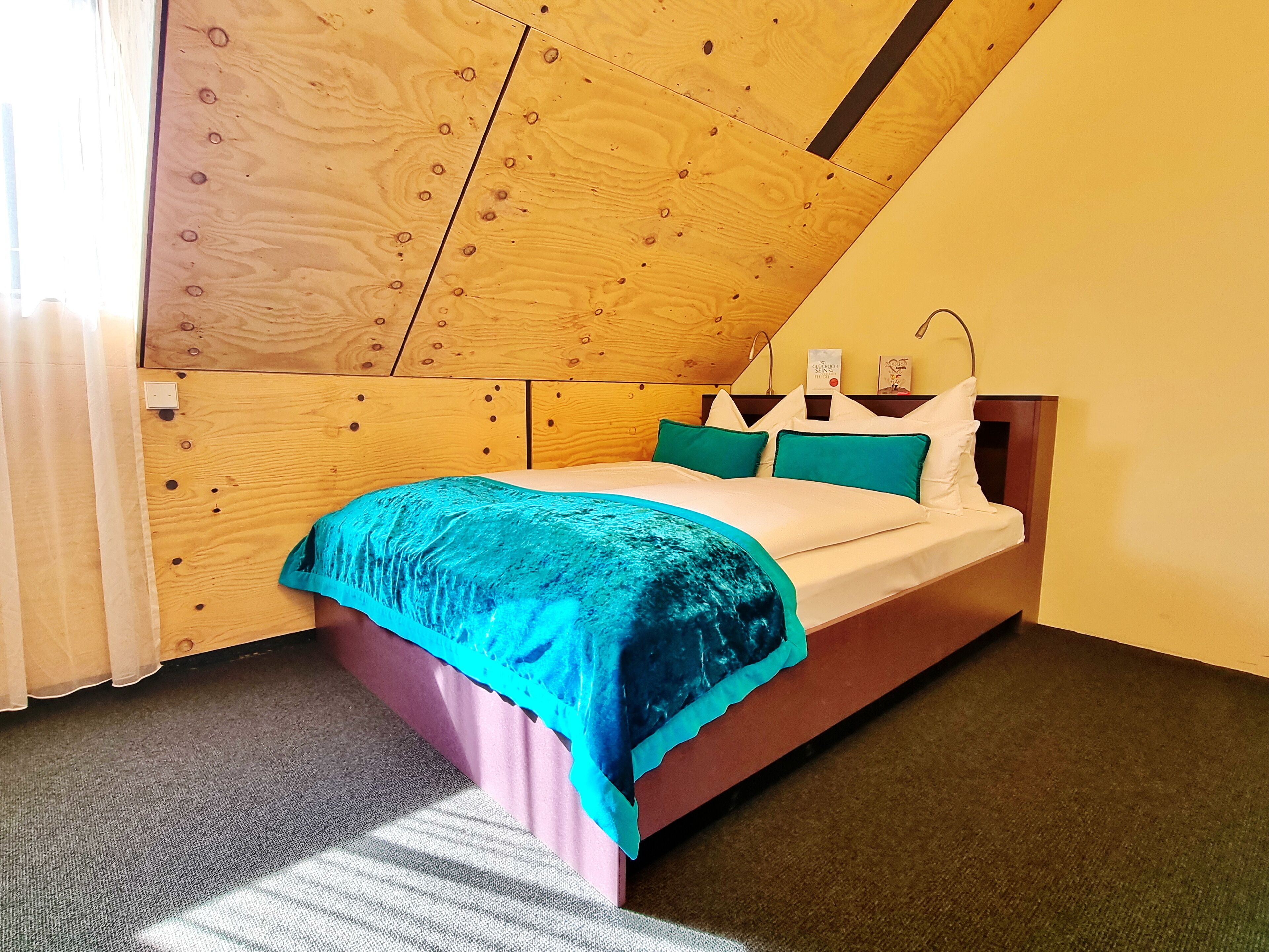A bedroom with a wooden sloped ceiling, a bed with teal and white bedding, and a nightstand with a lamp.