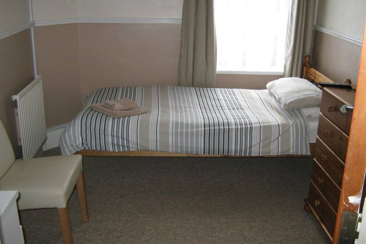 Une petite chambre avec un lit simple, une couverture à rayures, une commode en bois, une chaise et un radiateur.