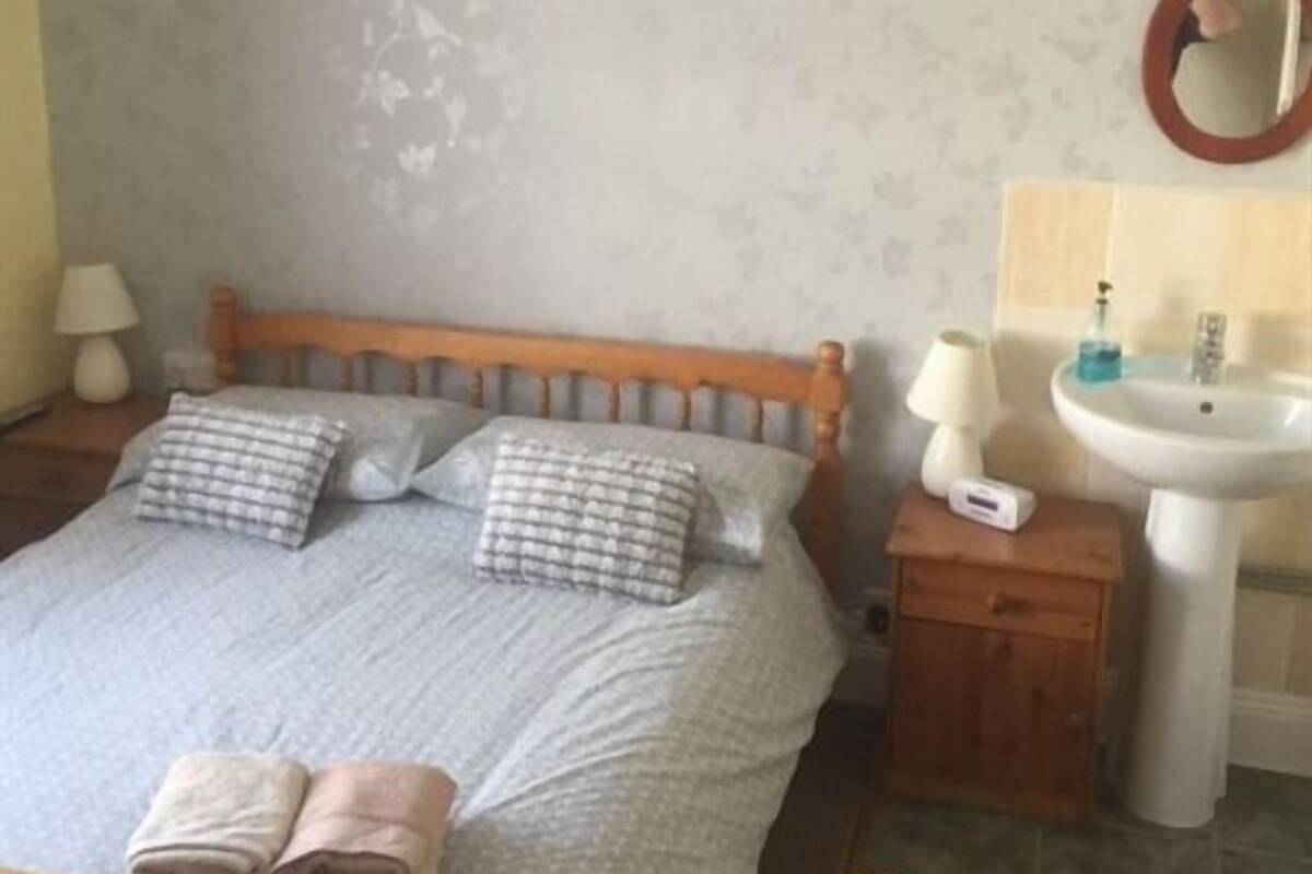 Une chambre à coucher avec un lit, des oreillers, une table de chevet avec une lampe, et une salle de bain avec un lavabo et un miroir.
