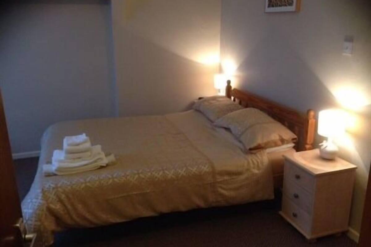Un lit bien fait, avec une couvre-lit beige et des oreillers, une table de chevet avec une lampe et une petite commode.