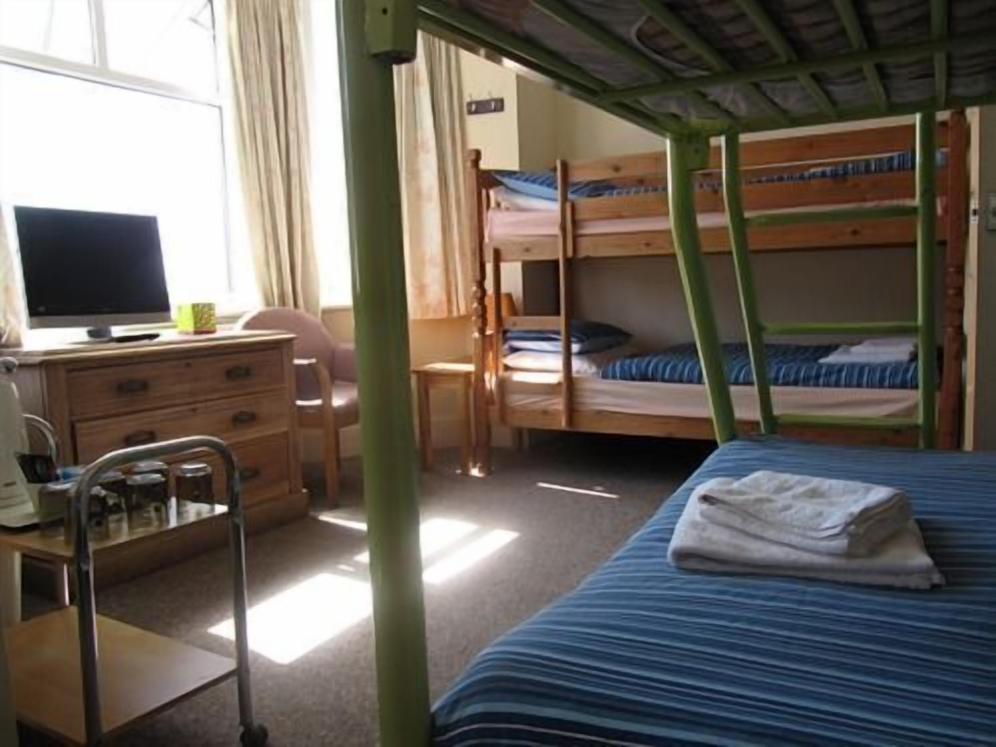 A dormitory room with bunk beds, a desk with a computer, a chair, and a small table with a lamp.