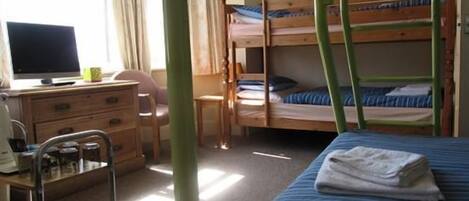 A dormitory room with bunk beds, a desk with a computer, a chair, and a small table with a lamp.
