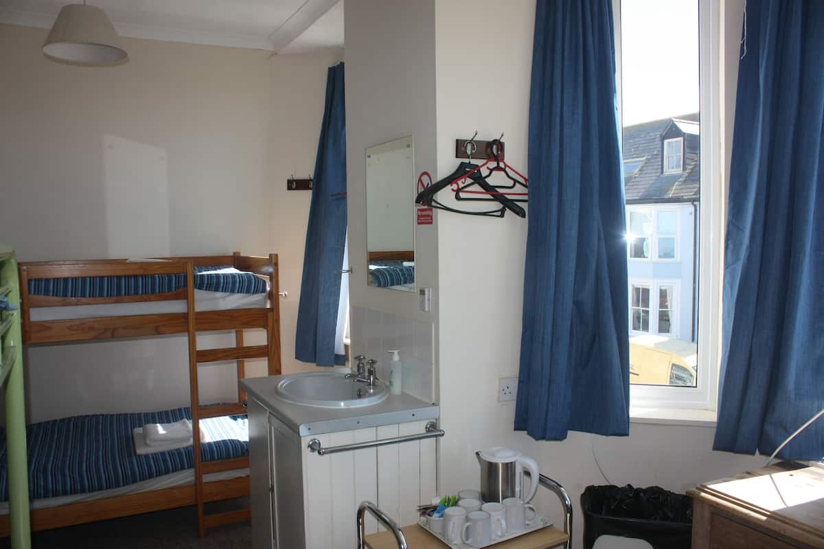 Habitación con literas, un lavabo y un espejo, una mesita con tazas, y vista a una casa a través de una ventana.