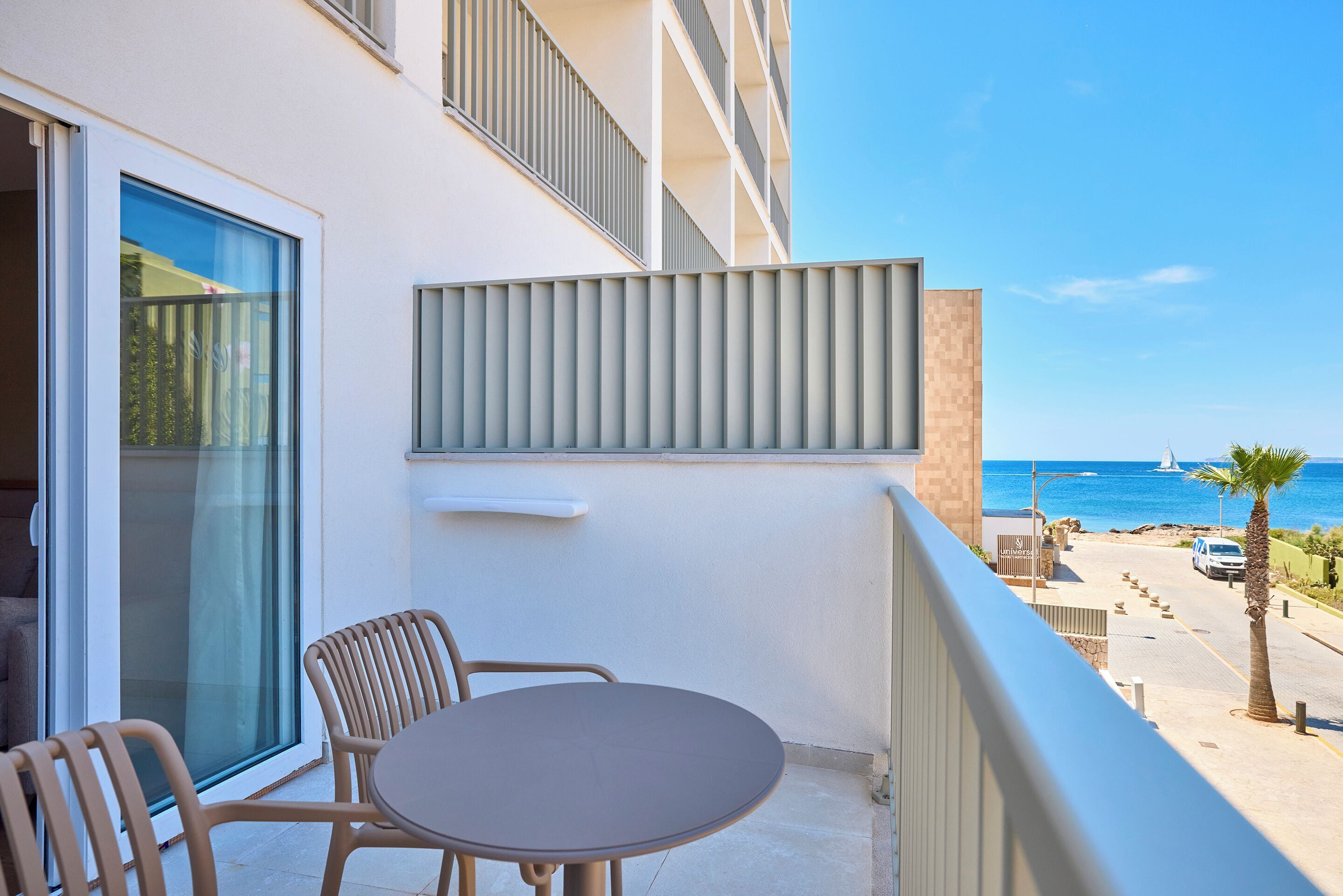 A balcony with a table and chairs, overlooking a coastal road with a palm tree and a building in the background.