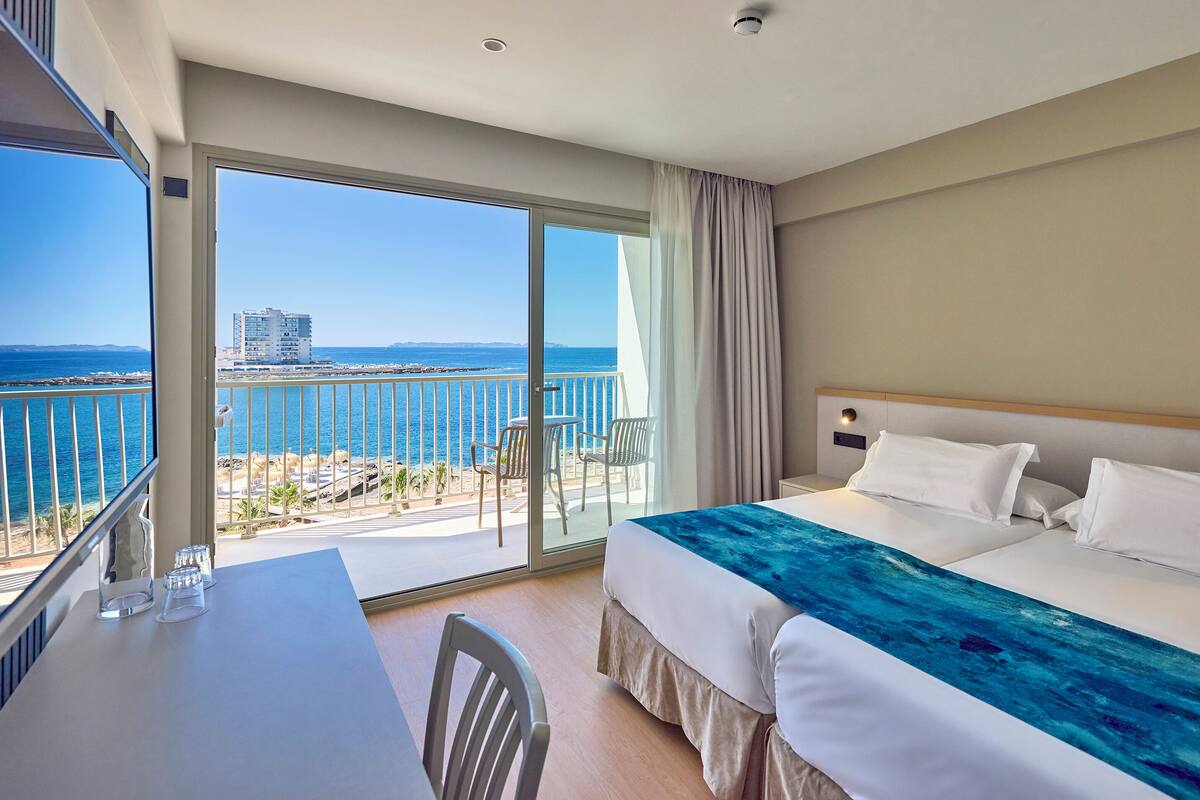 Quarto de hotel com varanda, cama com roupa de cama branca e vista para o mar e edifícios.