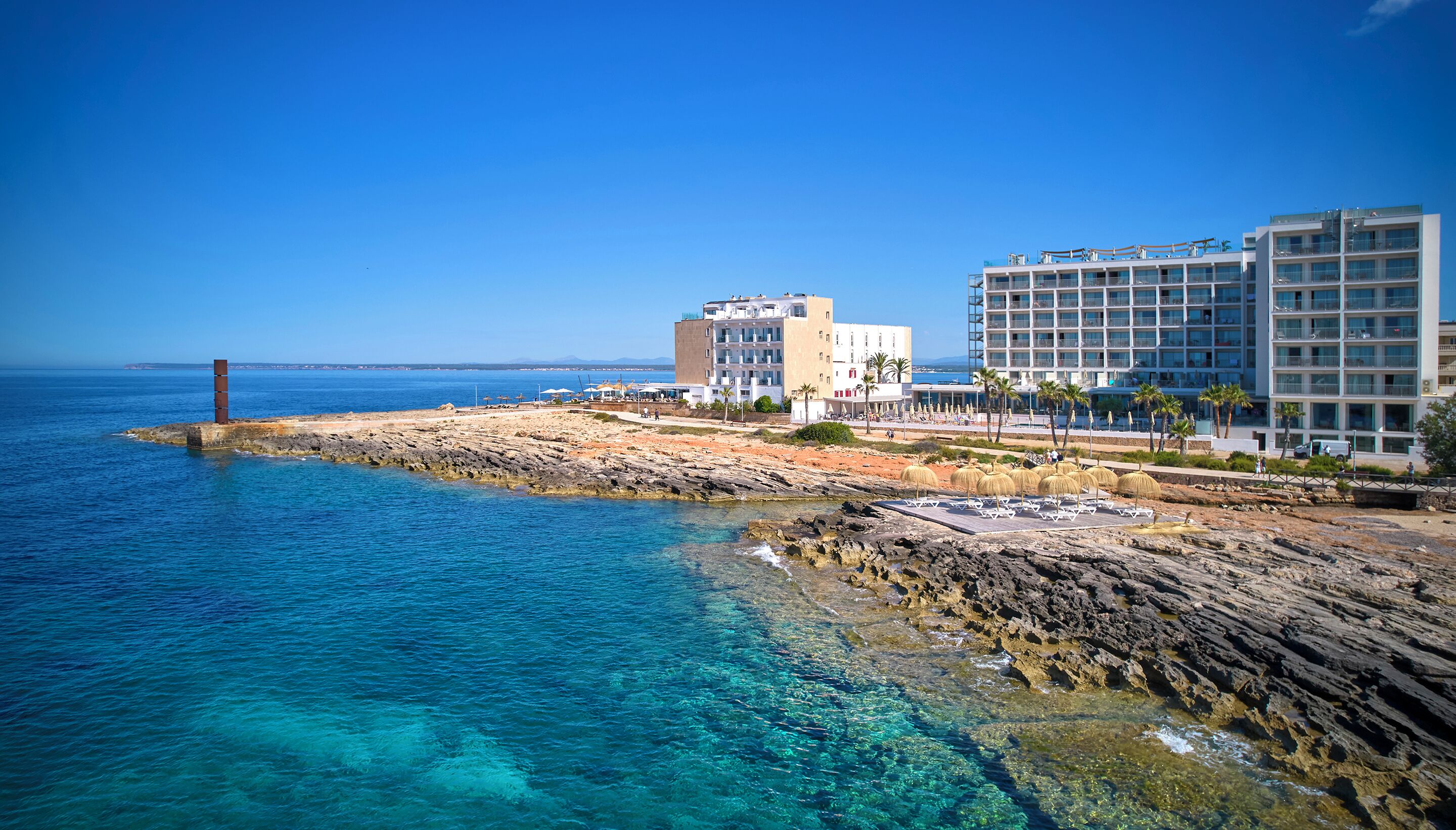 A coastal hotel with a rocky shoreline, clear blue waters, and modern buildings with balconies.