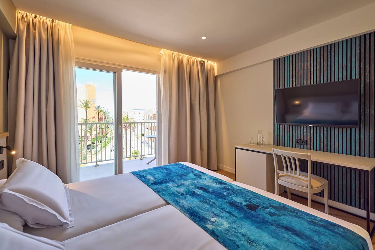Quarto de hotel com cama, escrivaninha, cadeira, televisão e varanda com cortinas.