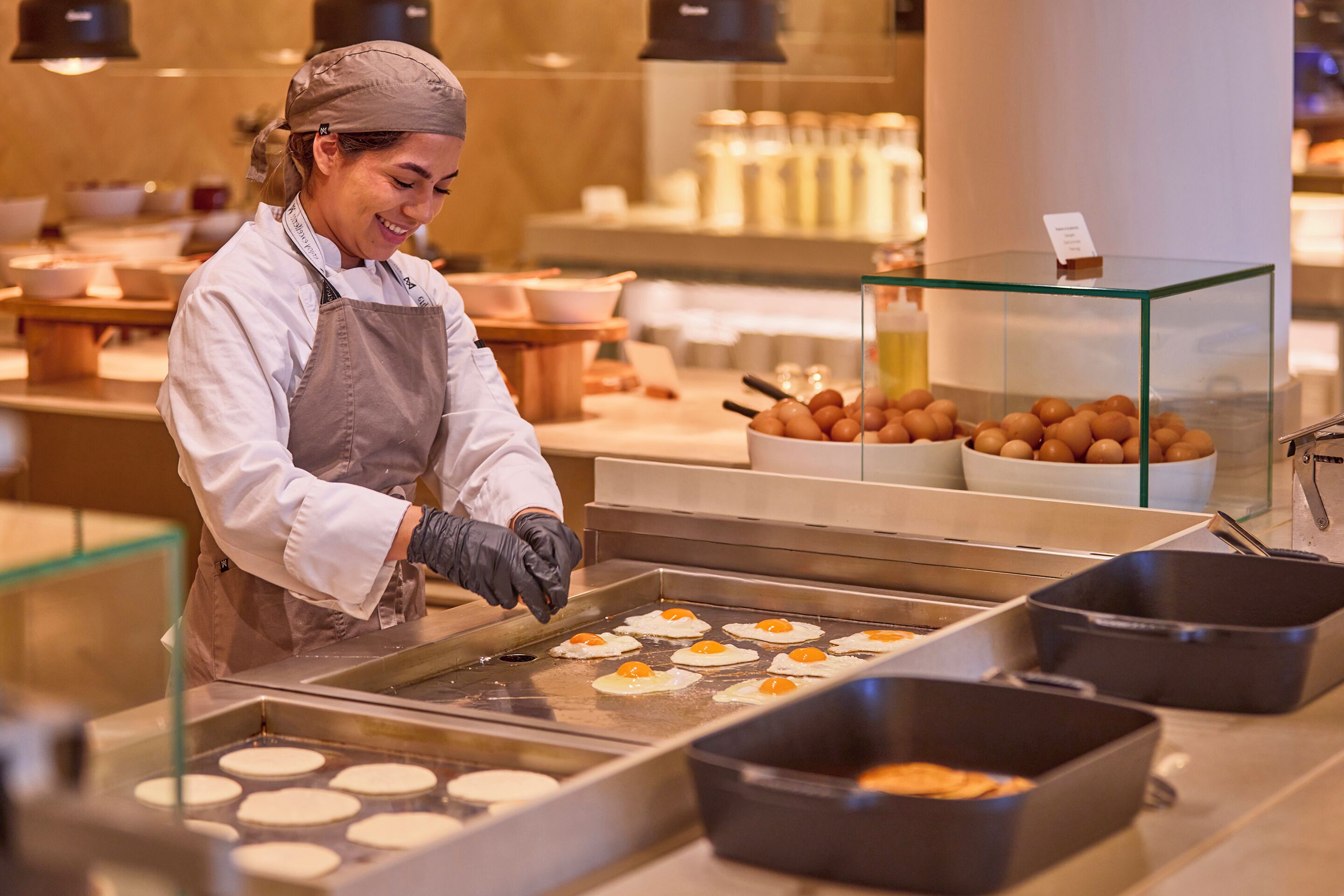 A person cooking at a commercial kitchen with multiple cooking stations and a display case with baked goods.