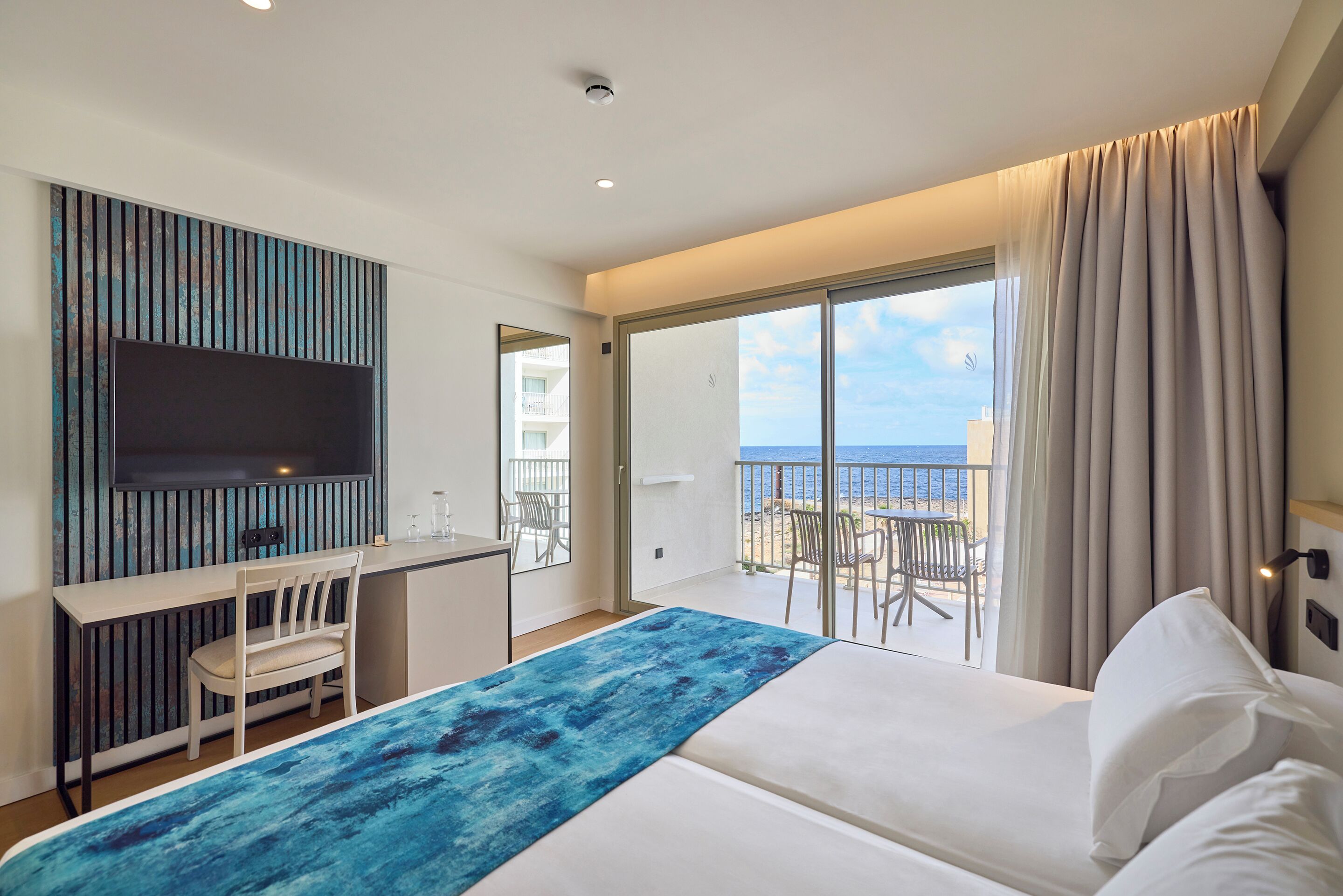 A hotel room with a bed, a desk, a chair, a TV, and a balcony with a view of the sea.