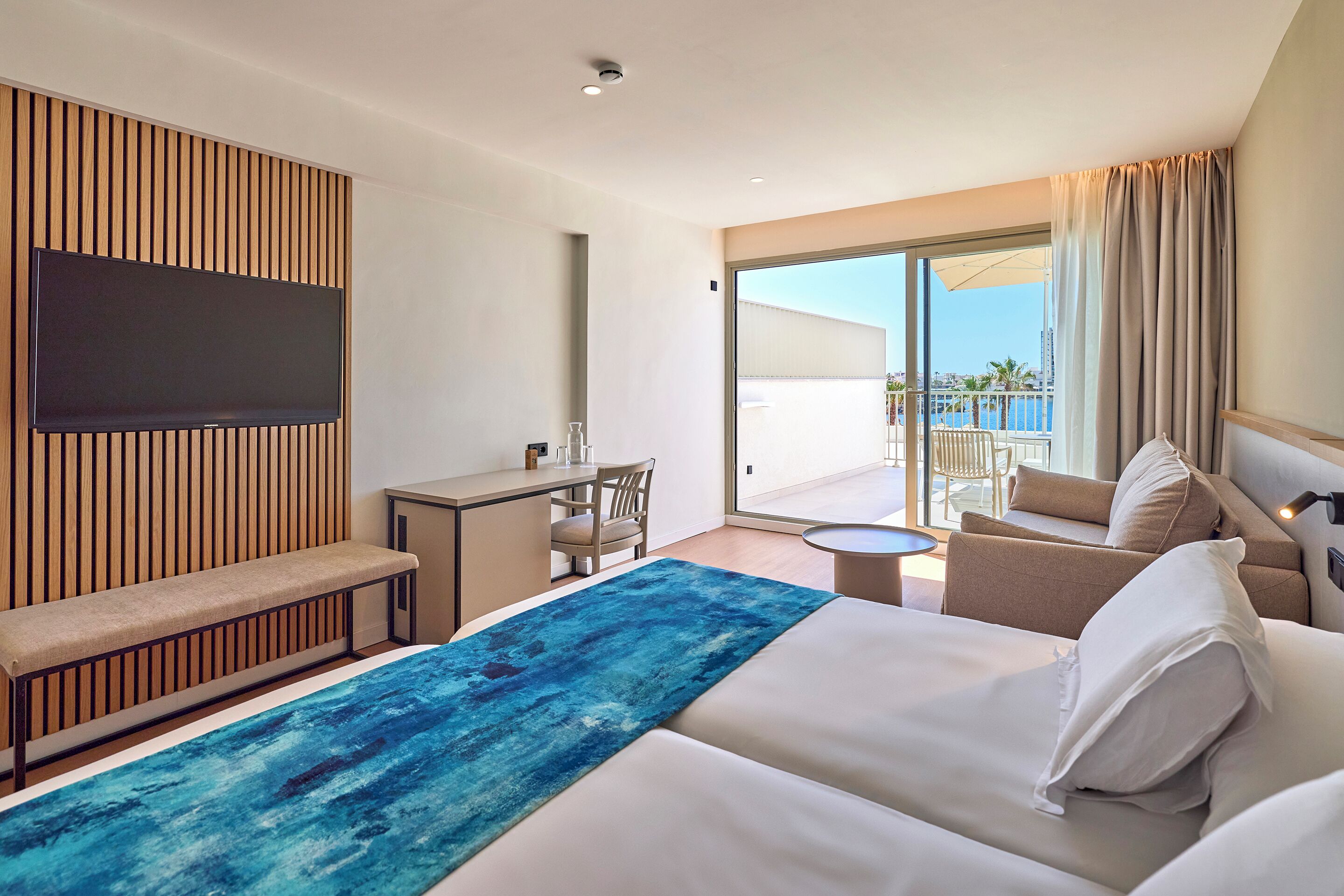 A hotel room with a large bed, a desk, a chair, a TV mounted on a wooden panel, and a balcony with a view.
