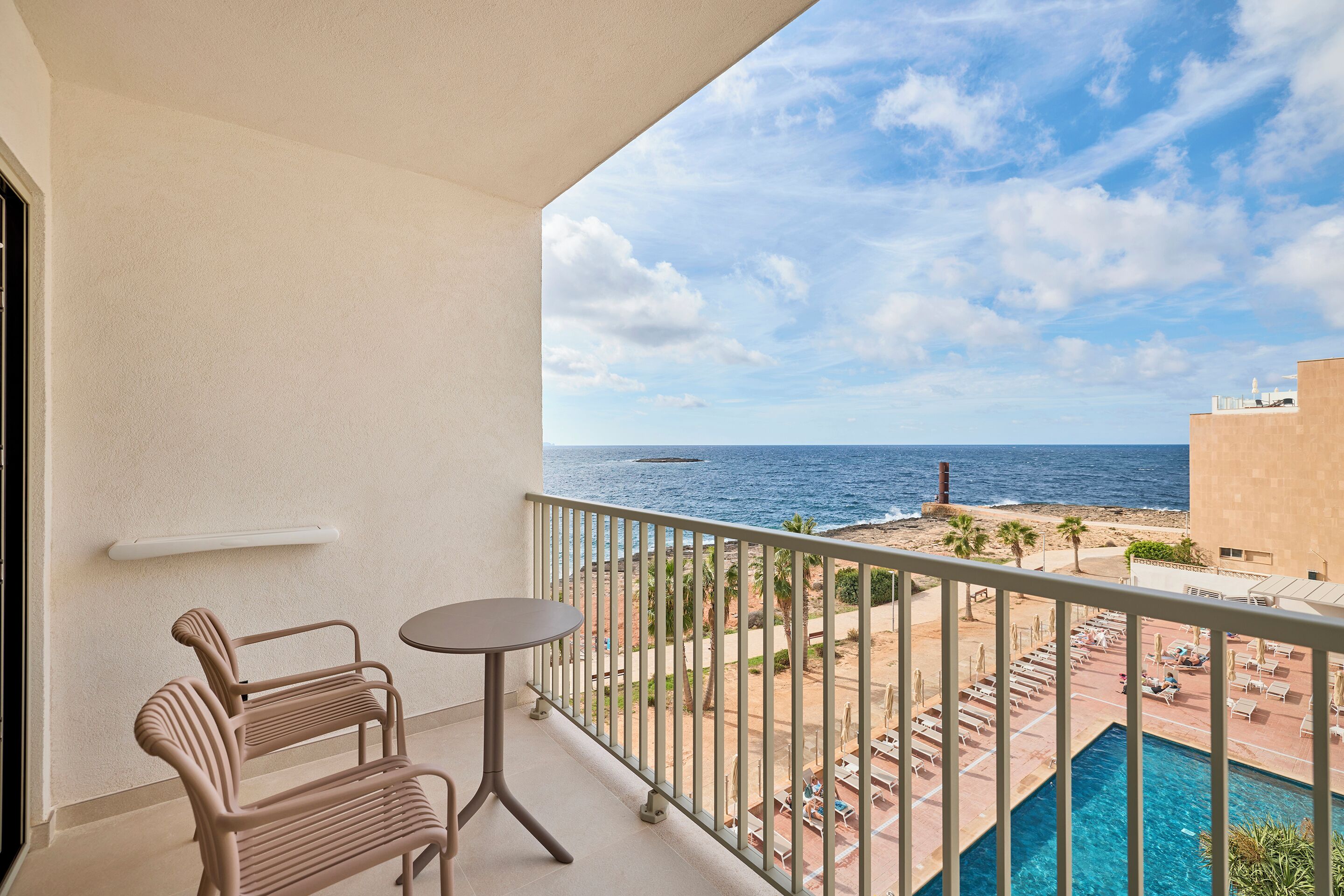 A balcony with a view of the sea, a pool, and lounge chairs.