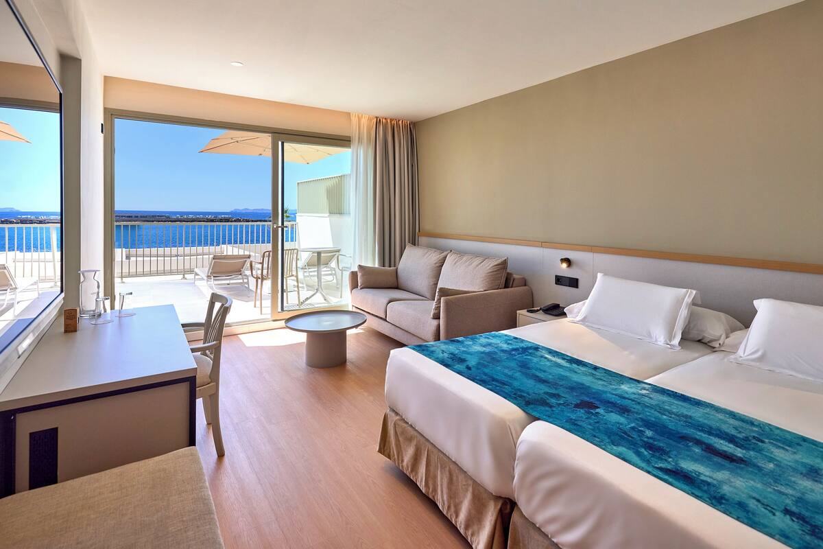 Quarto de hotel com varanda, uma cama, sofá, escrivaninha e cadeira.