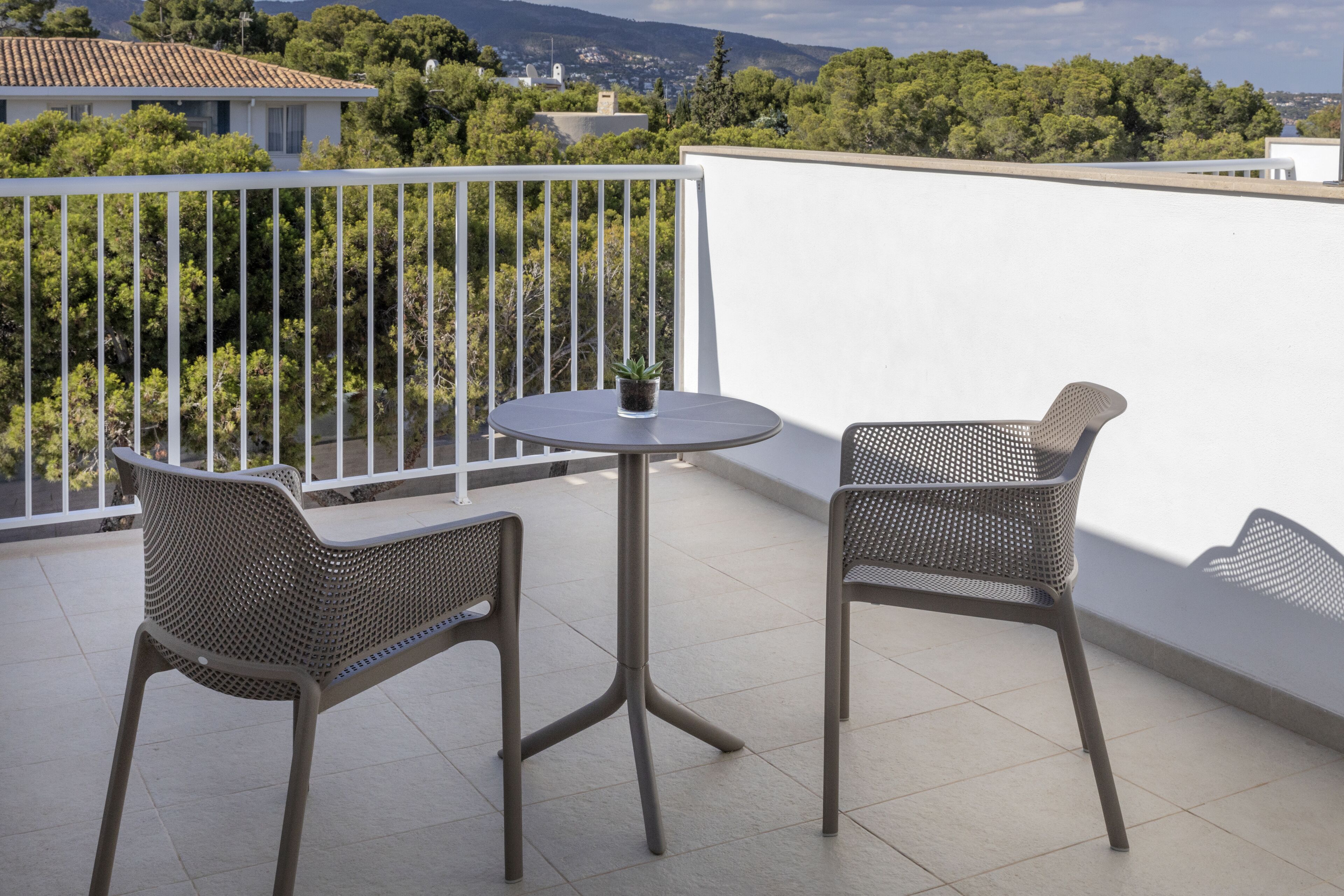 Un balcon avec deux fauteuils en osier et une petite table, d’où l’on peut admirer un paysage urbain avec des arbres et des bâtiments.