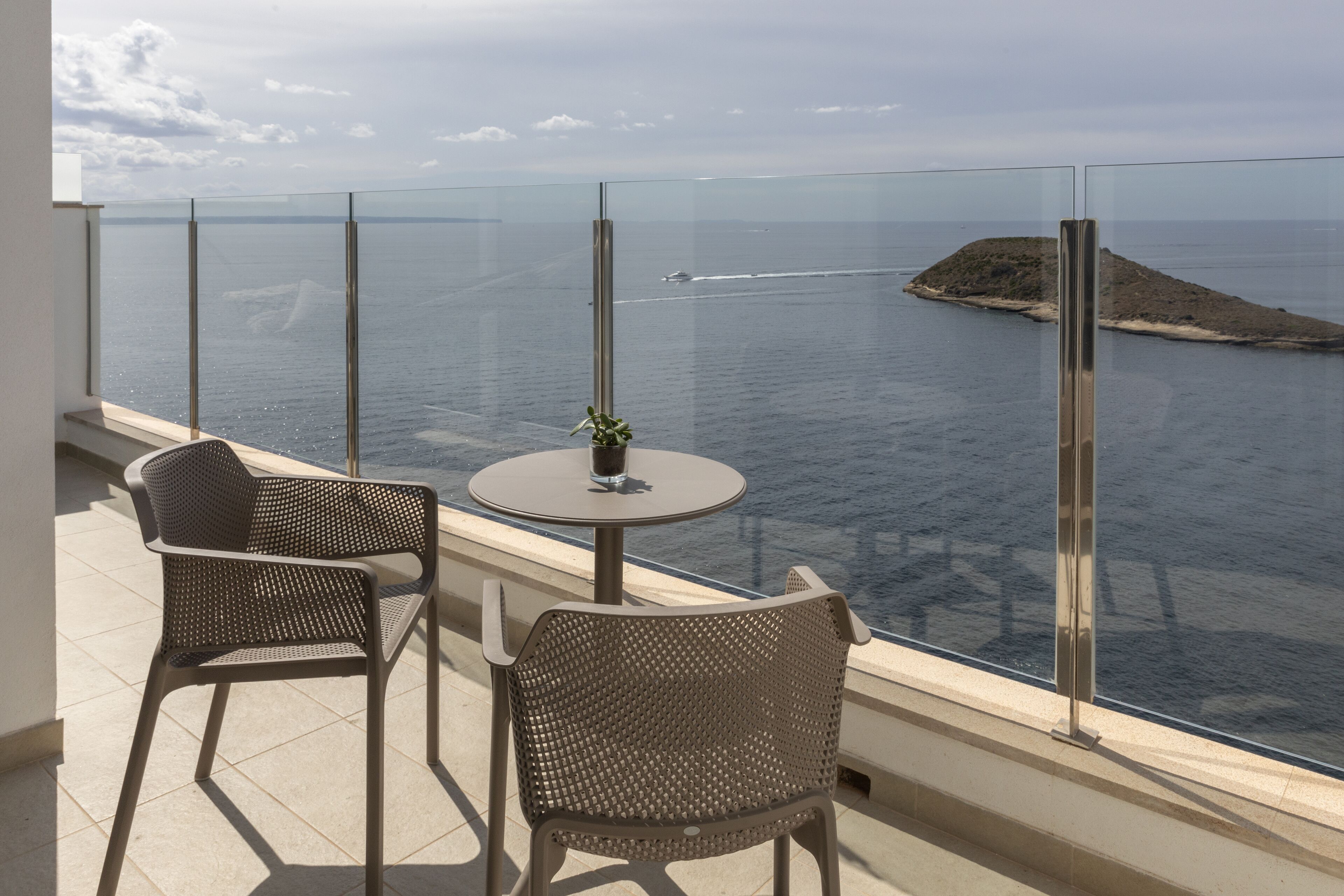Een balkon met twee rieten stoelen en een kleine ronde tafel, met uitzicht op een waterpartij en een eiland in de verte.