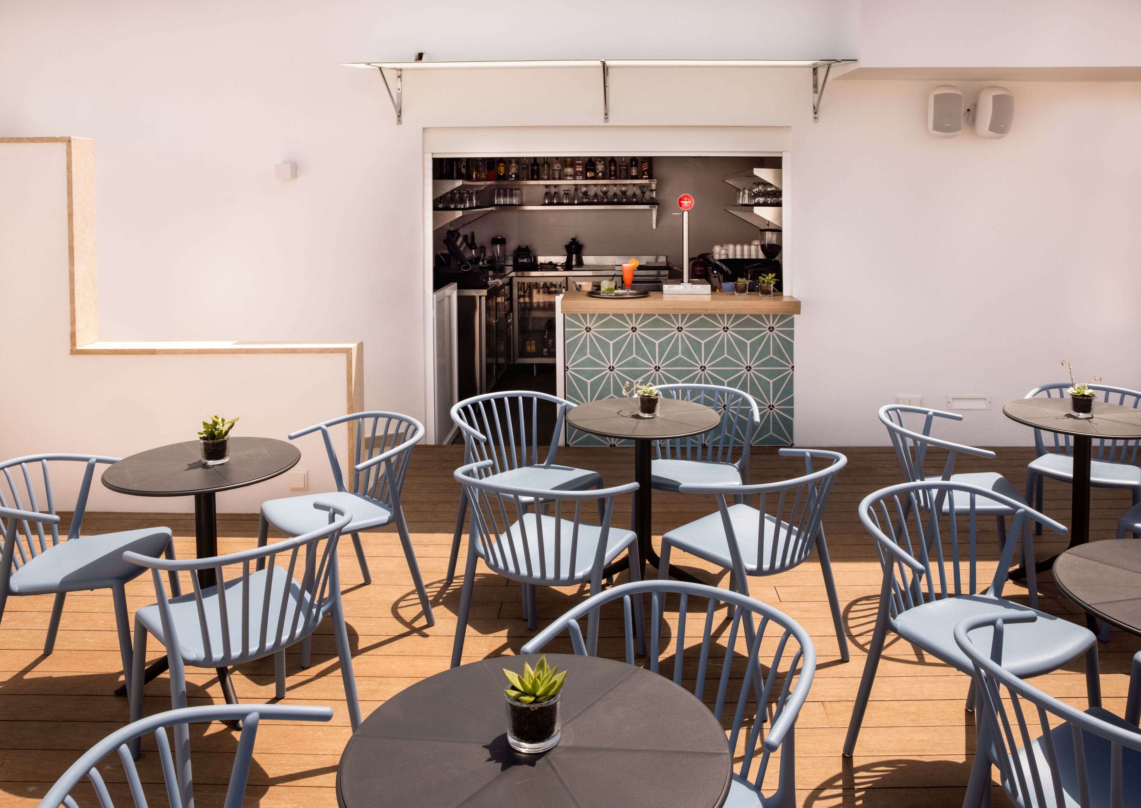 Een terras met ronde tafels en blauwe stoelen, een bar met flessenkasten, en uitzicht op de keuken door een open doorgang.