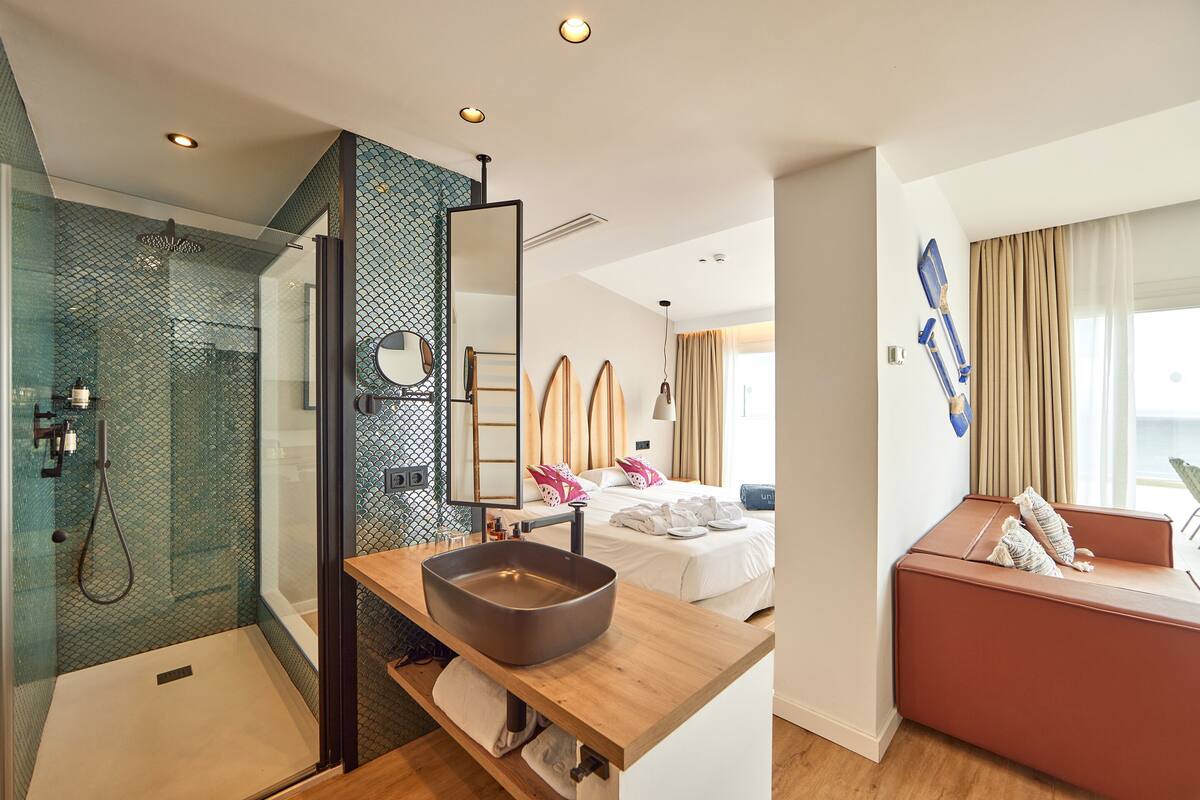 Une chambre d’hôtel moderne dotée d’une douche en verre, d’une coiffeuse en bois avec un grand lavabo, d’un lit avec du linge de maison blanc et d’un coin salon rouge offrant une vue sur la ville.