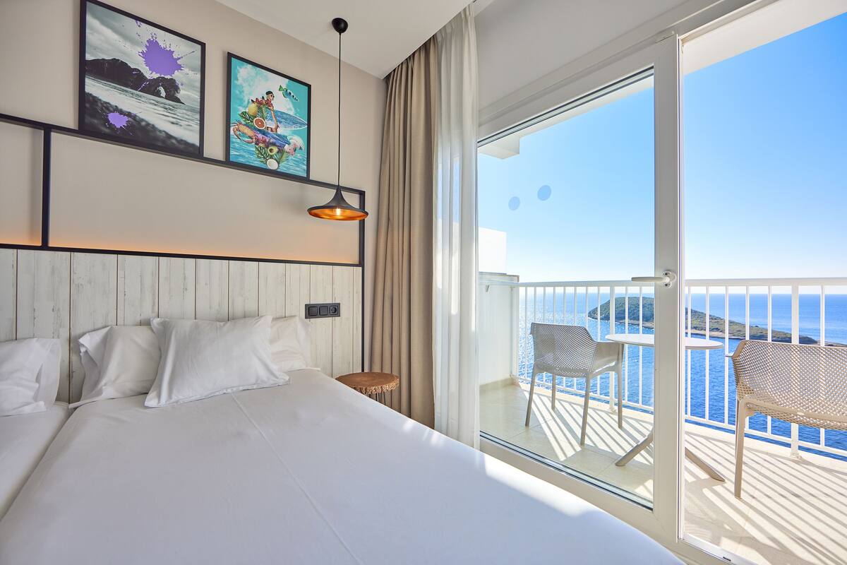 Une chambre d’hôtel avec un lit, une petite table, une chaise et un balcon donnant sur la mer.