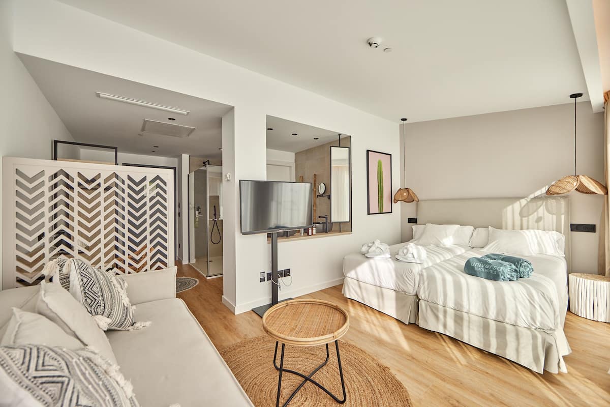 Une chambre moderne avec un grand lit, une télévision et un coin salon agrémenté d’un mur à motifs et d’un tabouret en osier.