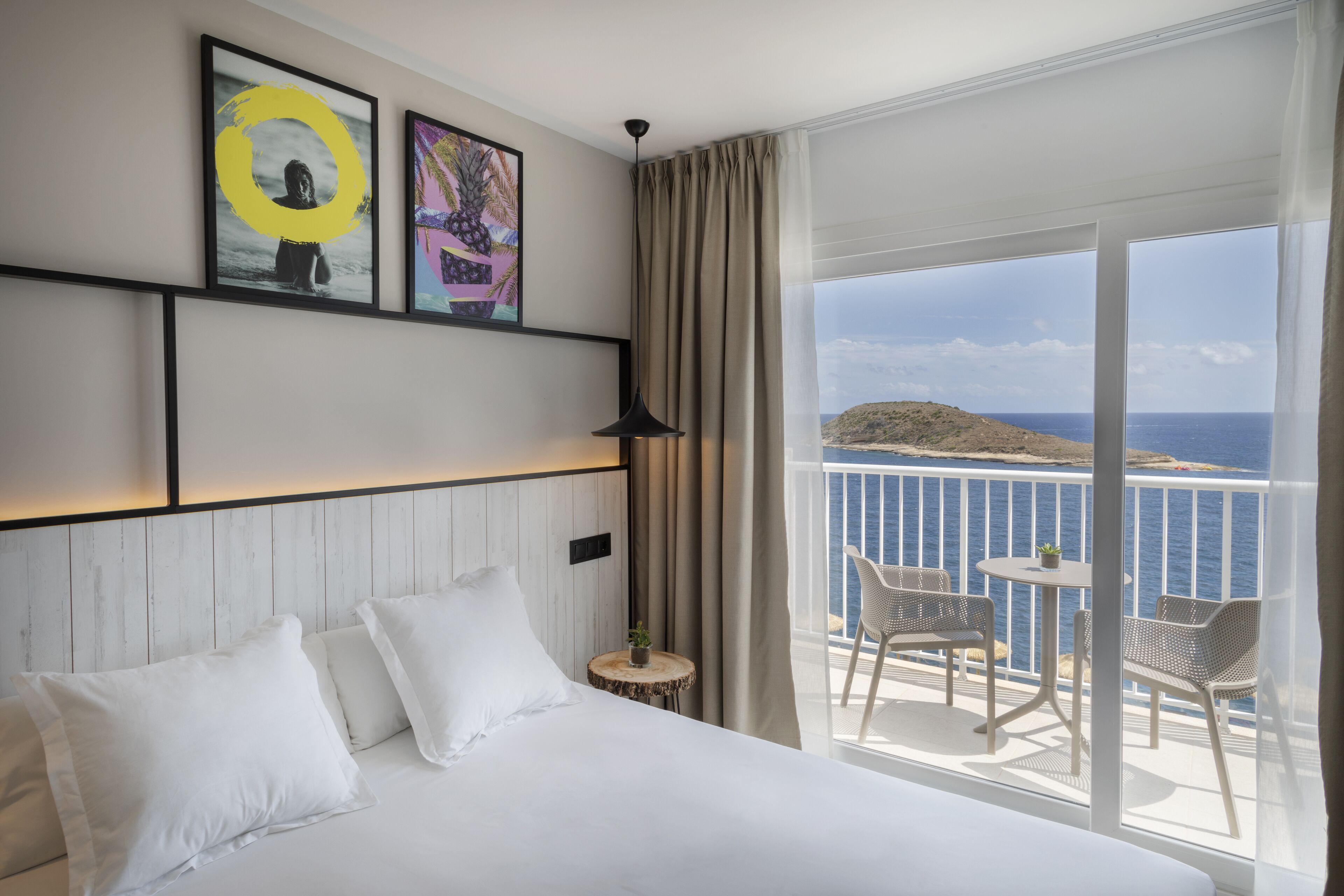 A hotel room with a large bed, a small round table with a plant, and a balcony with chairs and a view of the sea.