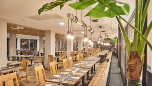 Un ampli restaurant amb taules i cadires de fusta allargades, llums penjants i plantes en testos.