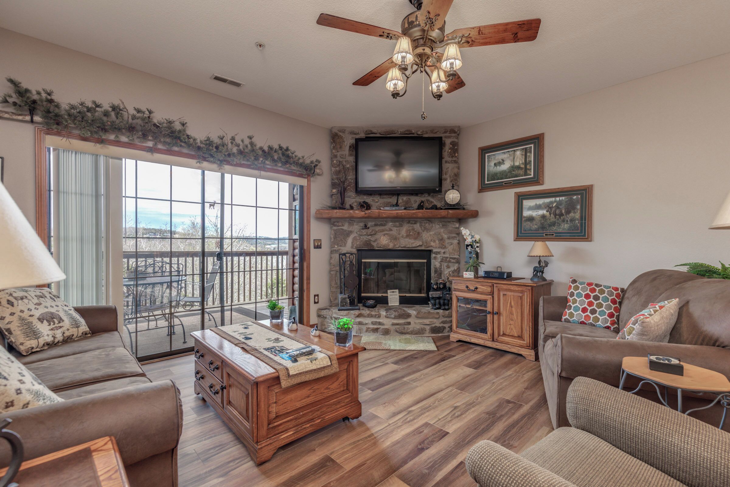 A spacious living room with a stone fireplace, a large TV mounted above it, and a ceiling fan.